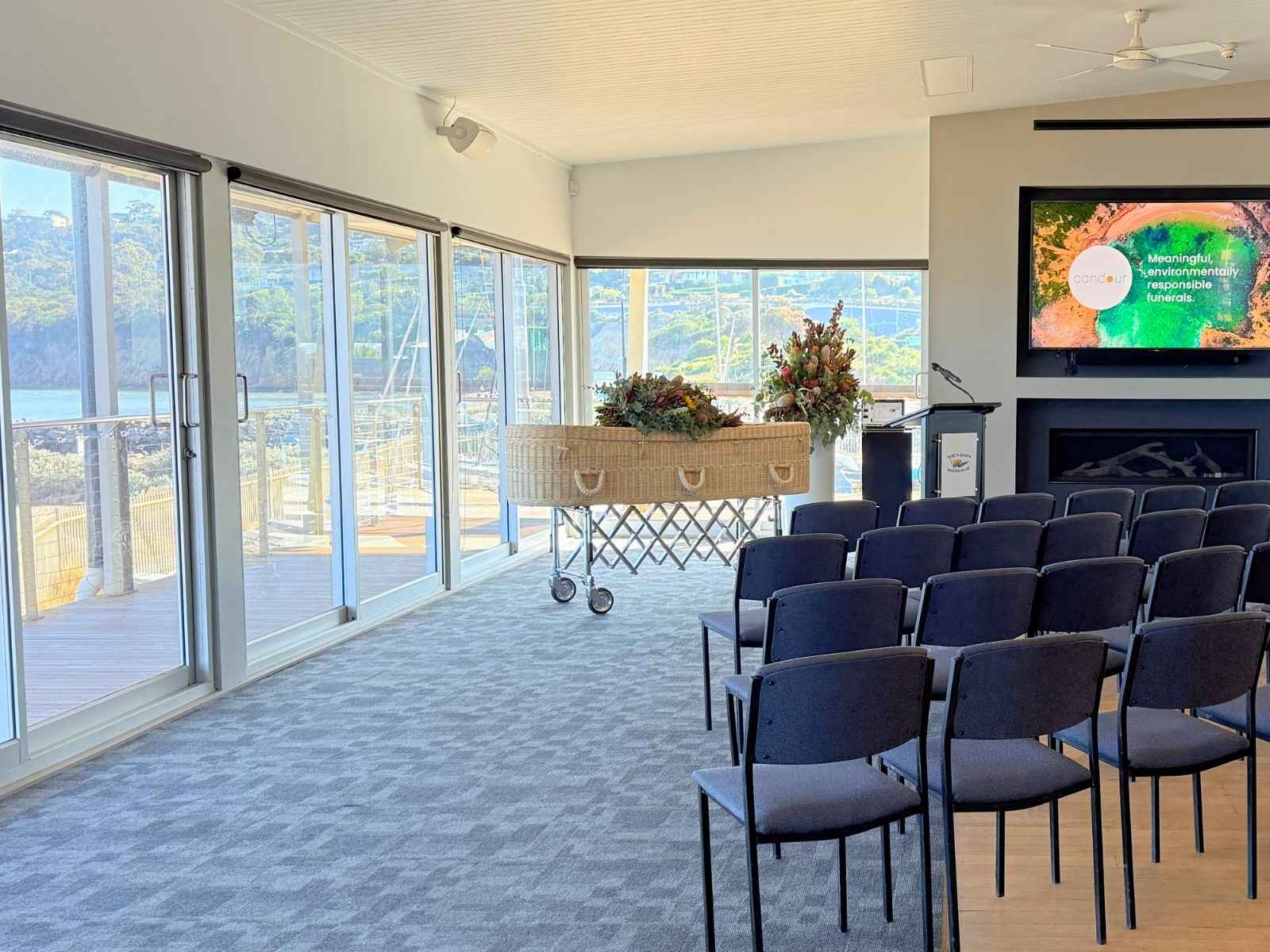 A bright and airy funeral venue with a podium, screen, and a floral-adorned coffin, surrounded by chairs and flooded with natural light from floor-to-ceiling windows overlooking the bay.
