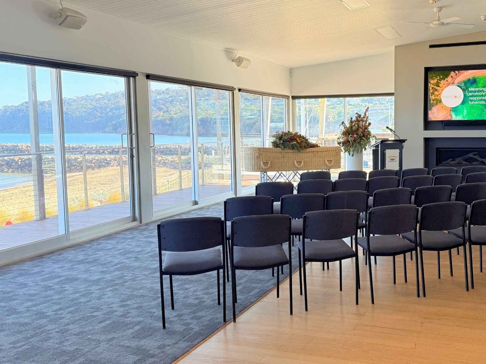 A spacious indoor setting with chairs arranged for a funeral service, offering sweeping views of Safety Beach and coastal cliffs through expansive glass sliding doors.