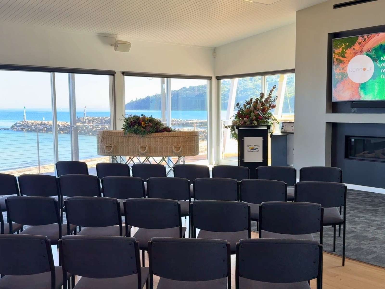A central view of a funeral room setup with a biodegradable coffin, elegant floral arrangements, a podium, and a fireplace beneath a large screen displaying a Candour Funerals message.