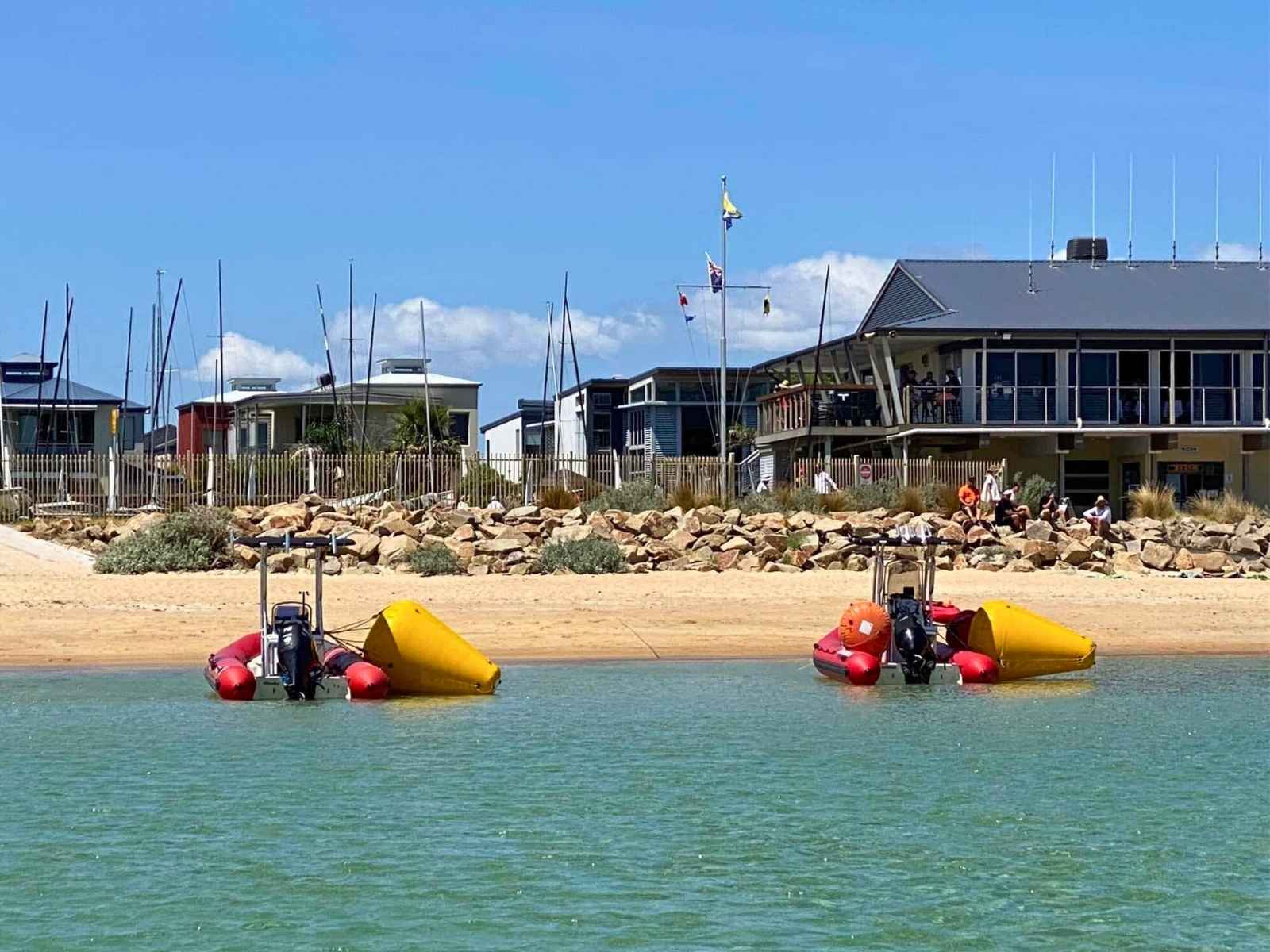The Safety Beach Sailing Club as seen from the water, with people gathered along the rocky shore, the modern building framed by sand, rocks, and colourful watercraft in the foreground.