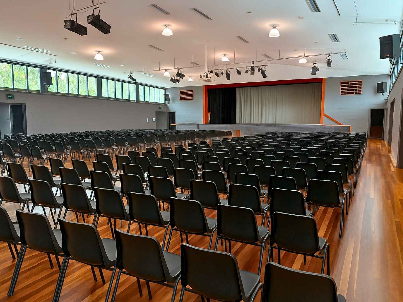 A large auditorium with hundreds of black chairs neatly arranged in rows, polished timber floors, and a stage with closed curtains and overhead theatrical lighting.