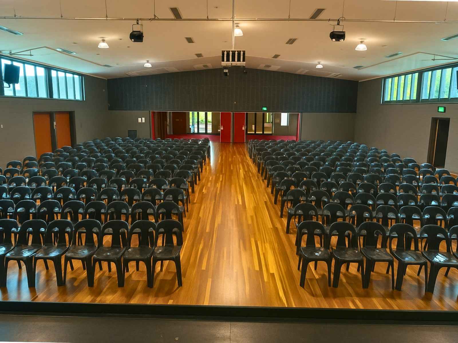 A view from the stage looking back across a full auditorium of black plastic chairs and polished wooden floors, leading to red entry doors at the far end of the hall.