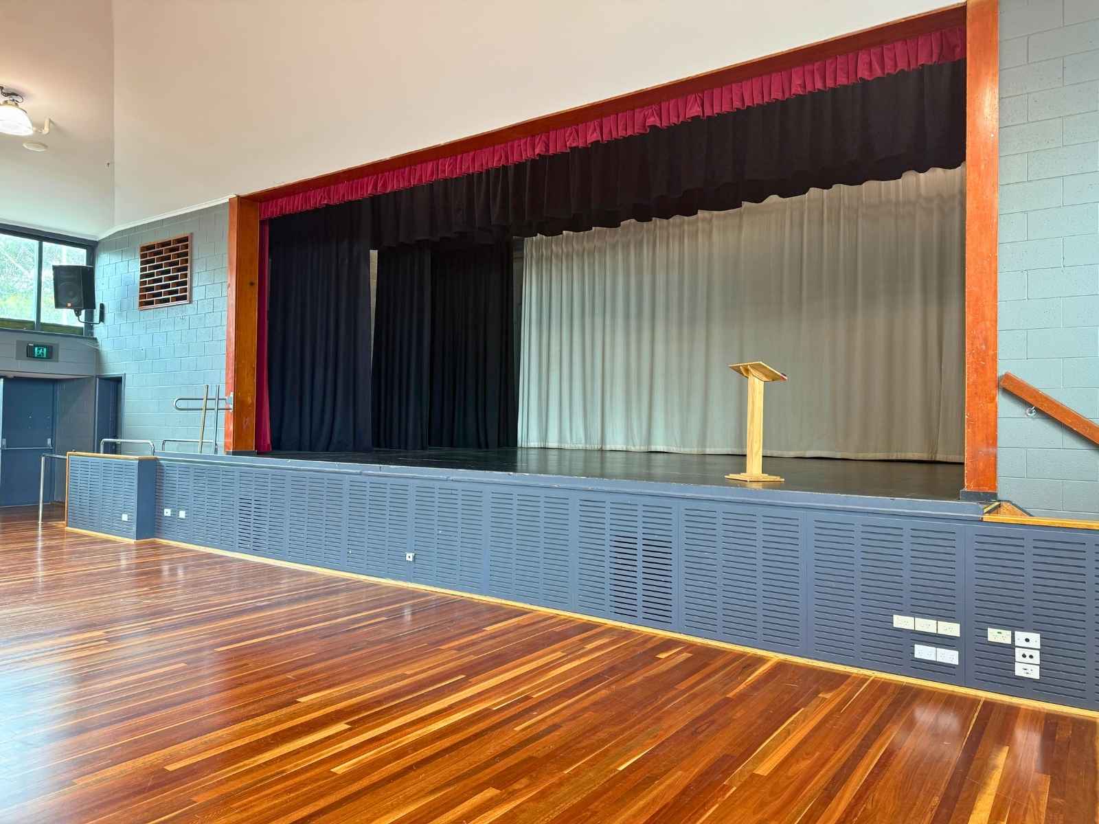 A close-up of the main stage at Peninsula Community Theatre featuring a simple wooden lectern, framed by red velvet curtains and surrounded by blue-grey walls.
