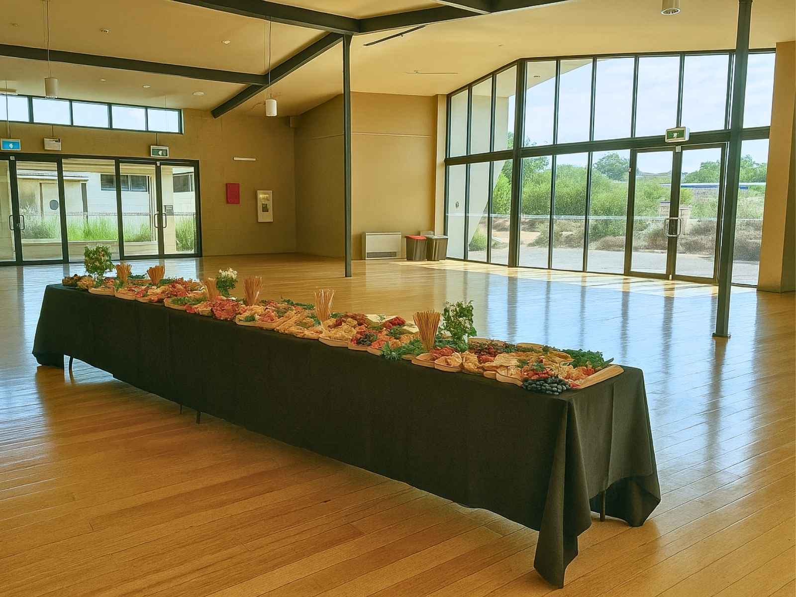 A bright, open reception area with floor-to-ceiling windows and a long grazing table laid with fresh produce and antipasto, set on polished timber floors.