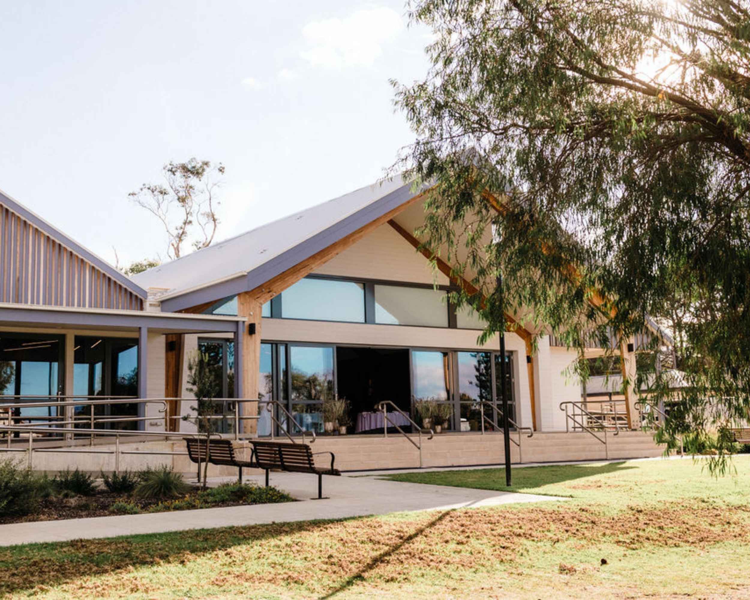 A contemporary, light-filled community building with pitched roofs, timber and glass detailing, and a wraparound ramp entrance, framed by native trees and soft afternoon light.