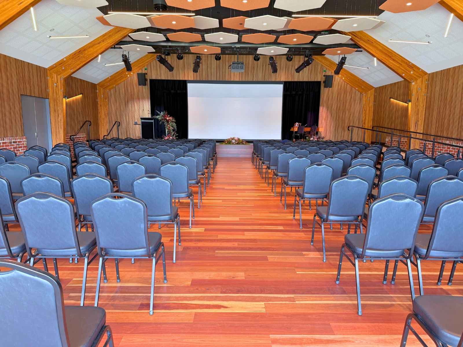 A modern timber-finished auditorium with rows of navy chairs arranged theatre-style, leading to a stage with screen and floral arrangements under a geometric acoustic ceiling.