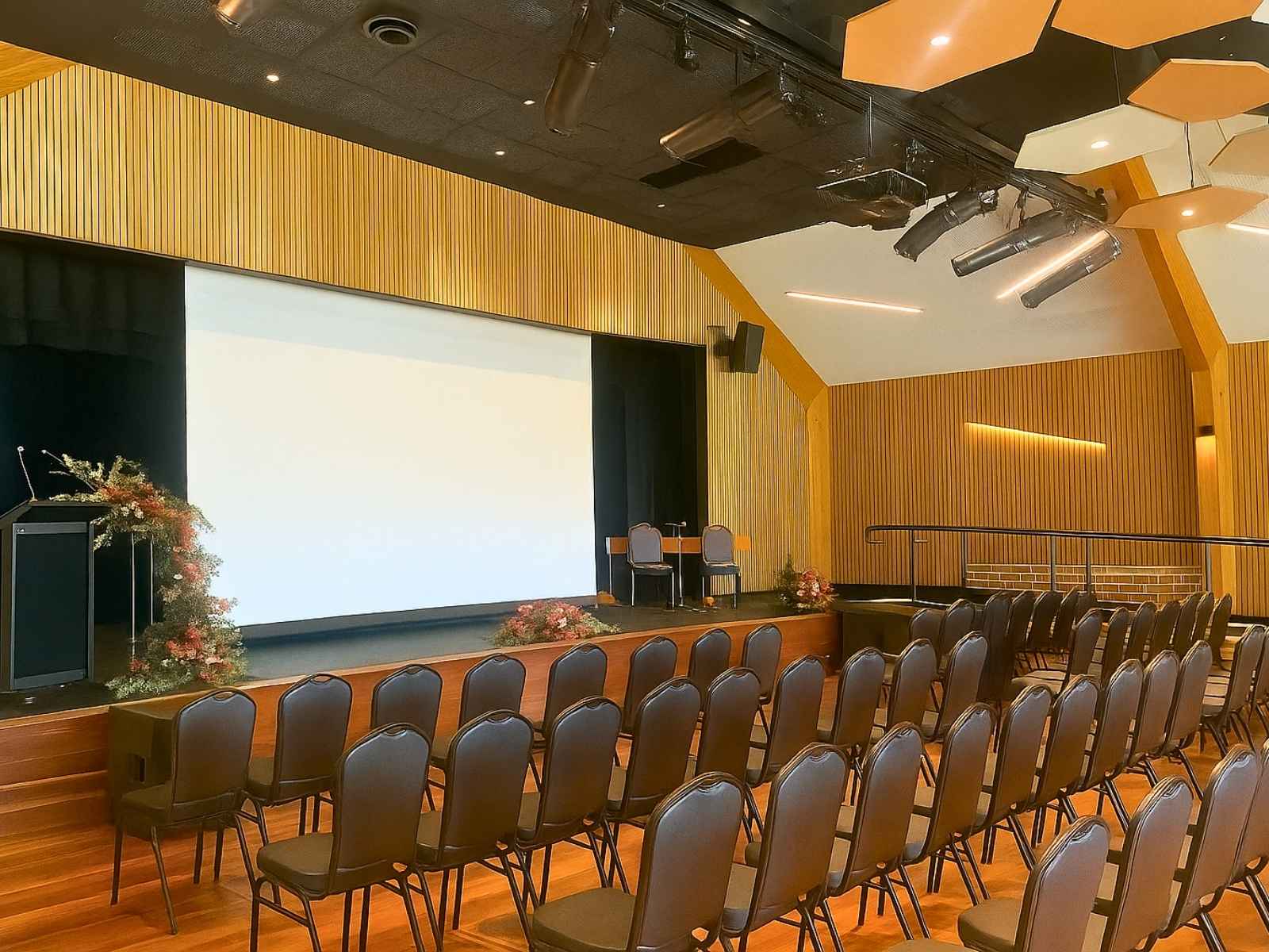 Angled view of the Flinders Civic Hall stage with dark seating, floral tributes, and a large projector screen, framed by vertical timber paneling and architectural lighting.