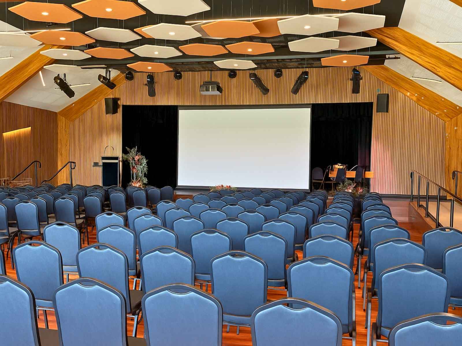 Straight-on perspective down the aisle of a spacious civic hall, showing organised seating, timber finishes, and a large projection screen beneath hanging lights and acoustic panels.