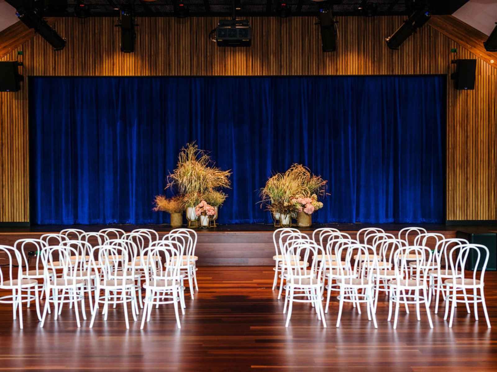 A vibrant and theatrical setting featuring white bentwood chairs arranged before a deep blue curtain and decorative native floral arrangements on the stage.