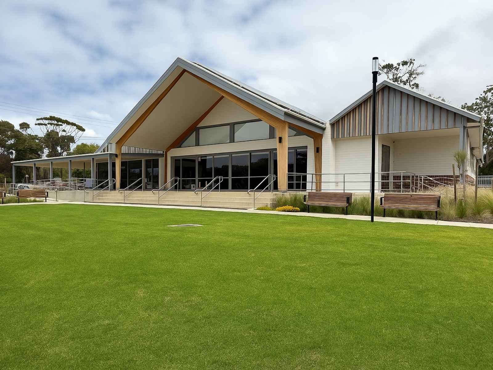 A spacious, modern civic hall with timber and white finishes, viewed from across a perfectly manicured lawn, with accessible ramp entryways and timber benches lining the path.