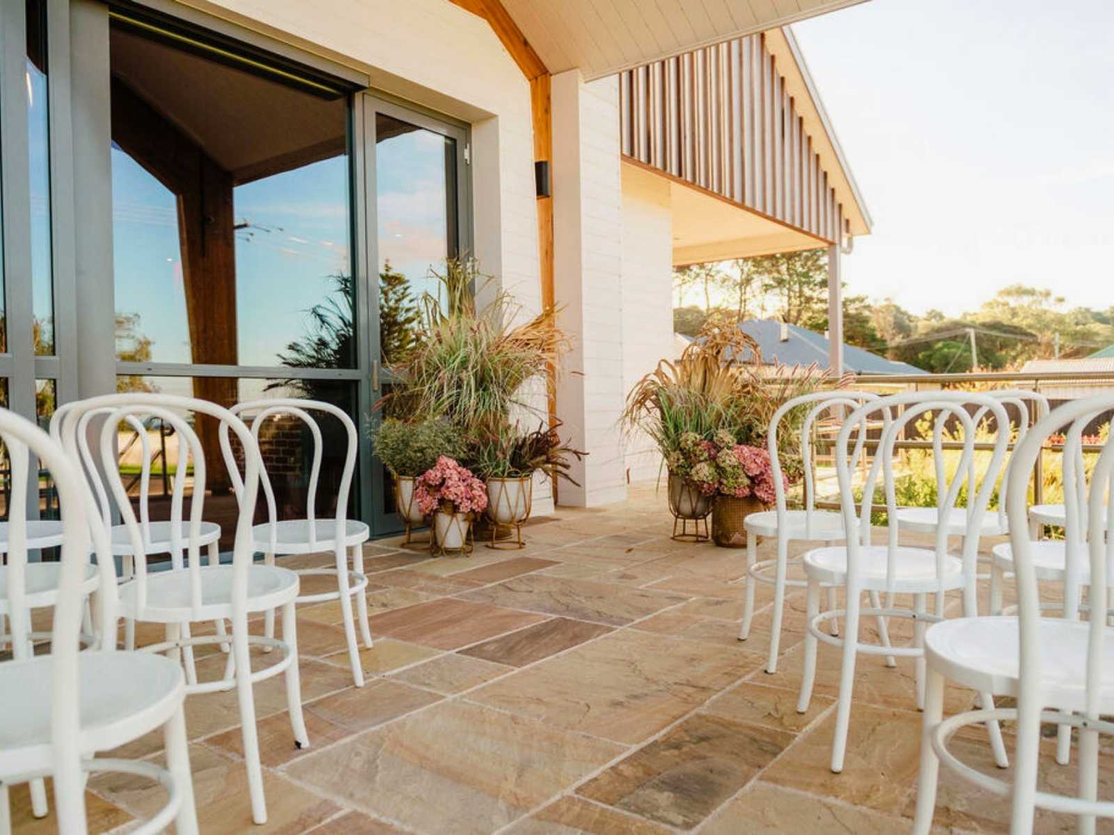A charming outdoor setting at Flinders Civic Hall featuring white bentwood chairs arranged on a stone patio, surrounded by lush floral arrangements and soft natural light.