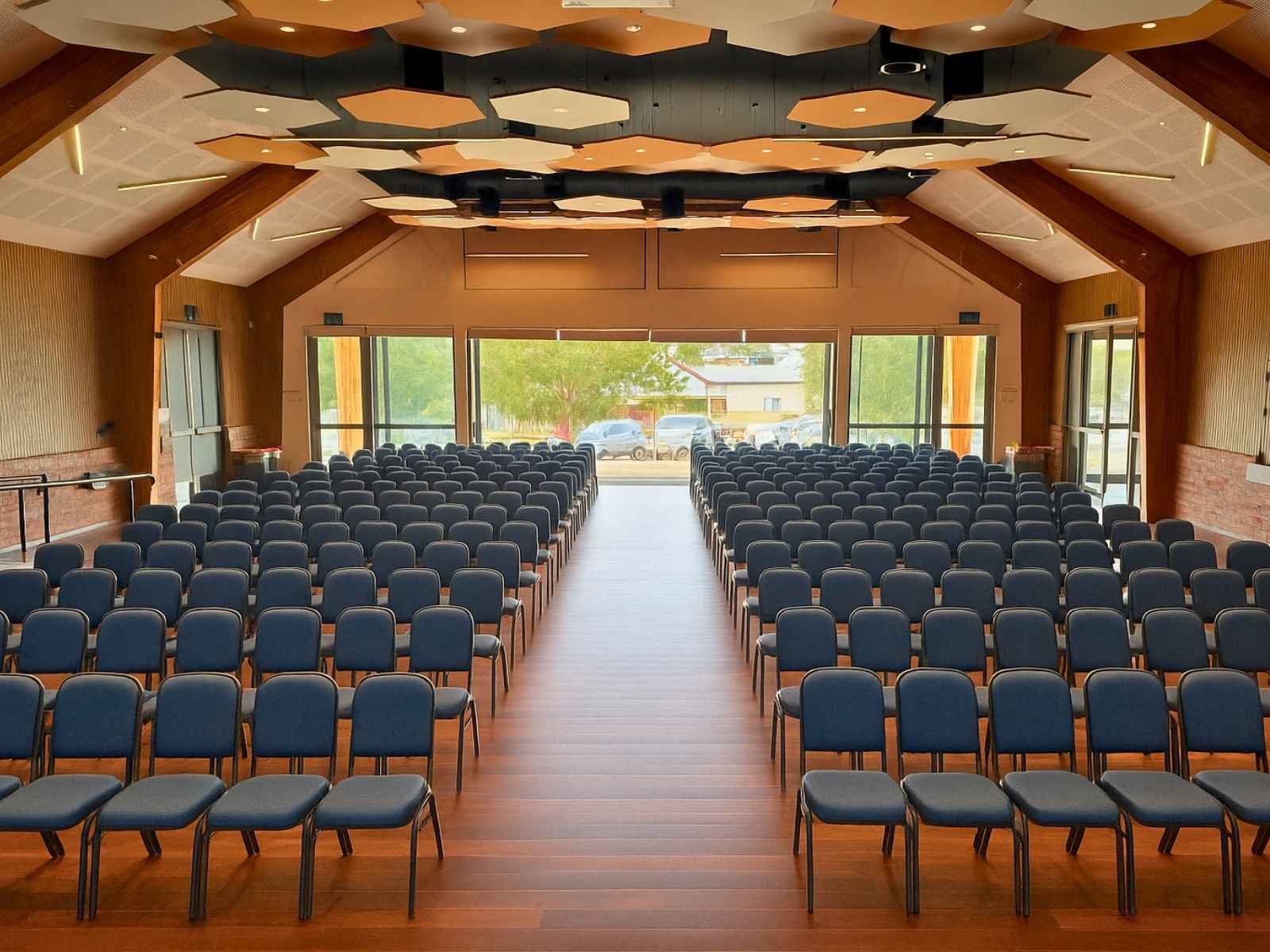A wide auditorium view from the stage looking back towards large windows and glass doors, with rows of chairs and architectural ceiling panels visible above.