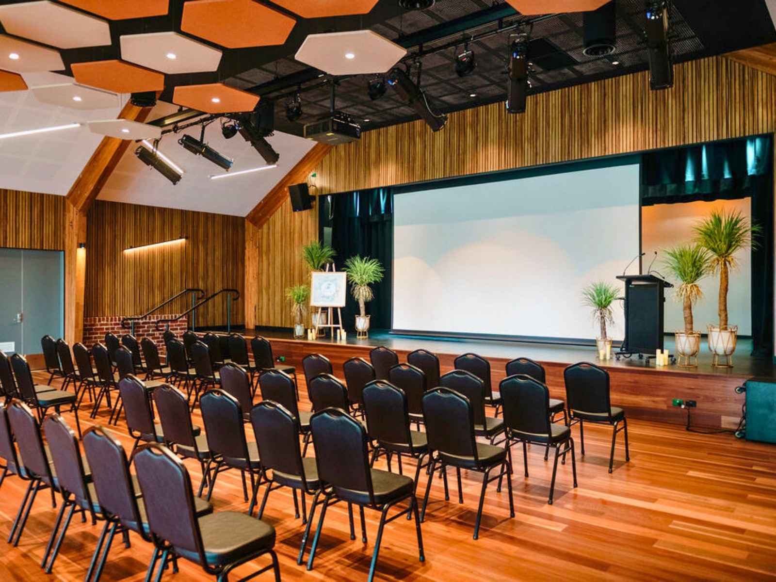 A warm and modern civic space with black chairs, lush potted plants, and a framed display on an easel, set against timber panelled walls and a large projection screen.