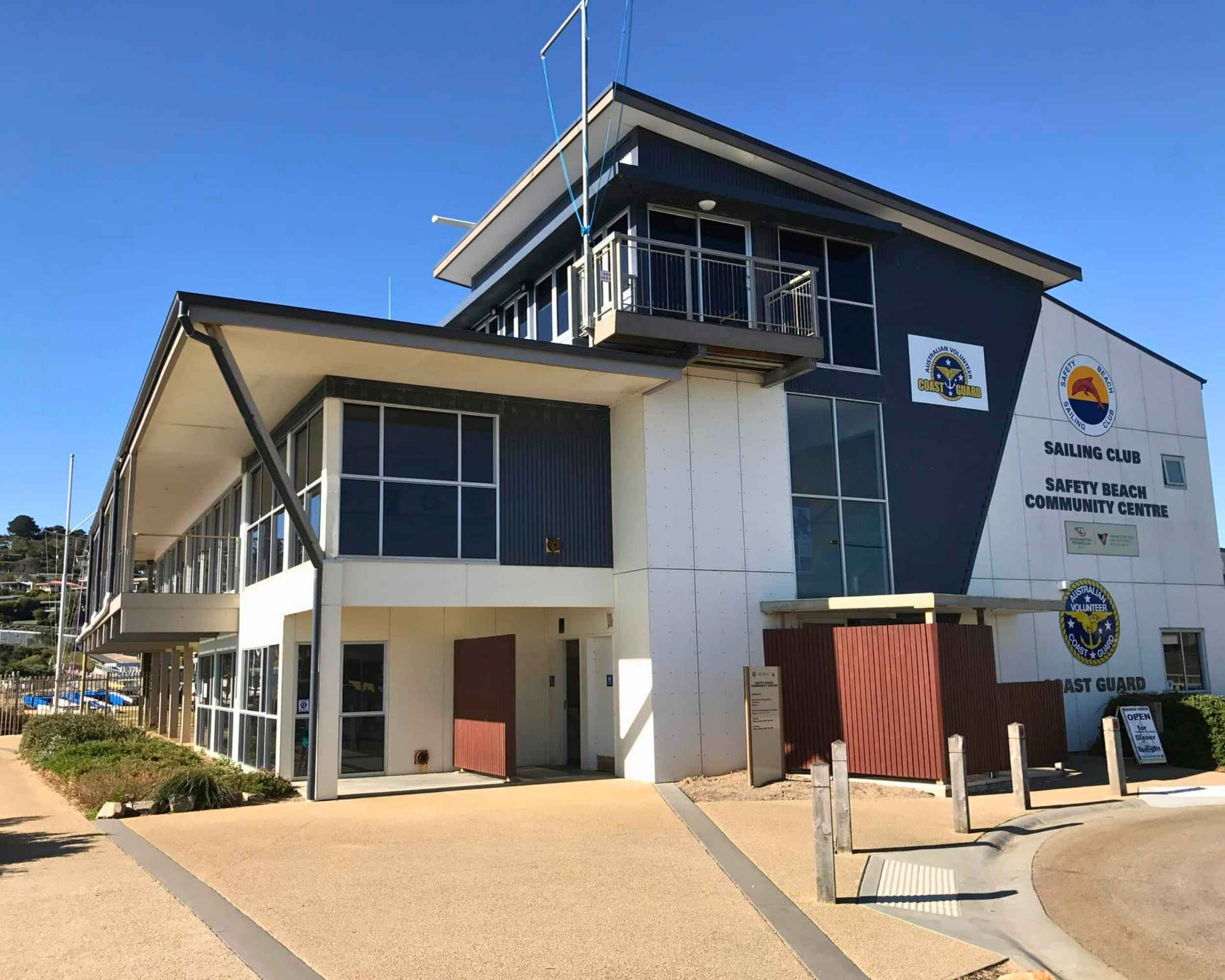 The exterior of the Safety Beach Sailing Club and Community Centre, a modern two-storey building with clean lines and nautical signage, located right by the coast under a clear blue sky.