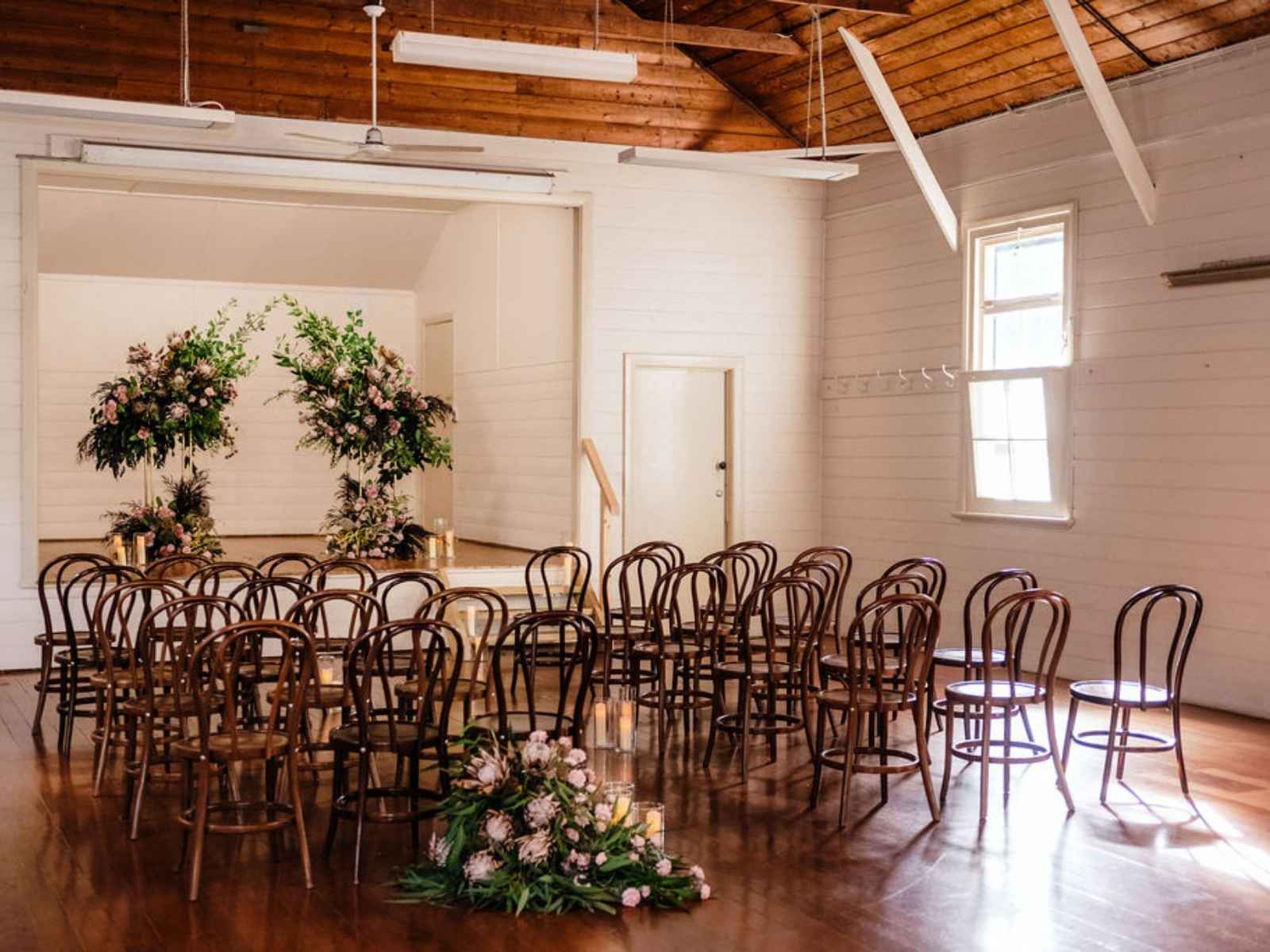 A charming timber hall with white-painted walls and exposed wood ceiling, set for a memorial with dark wooden chairs, lush floral arrangements, and glowing candles.