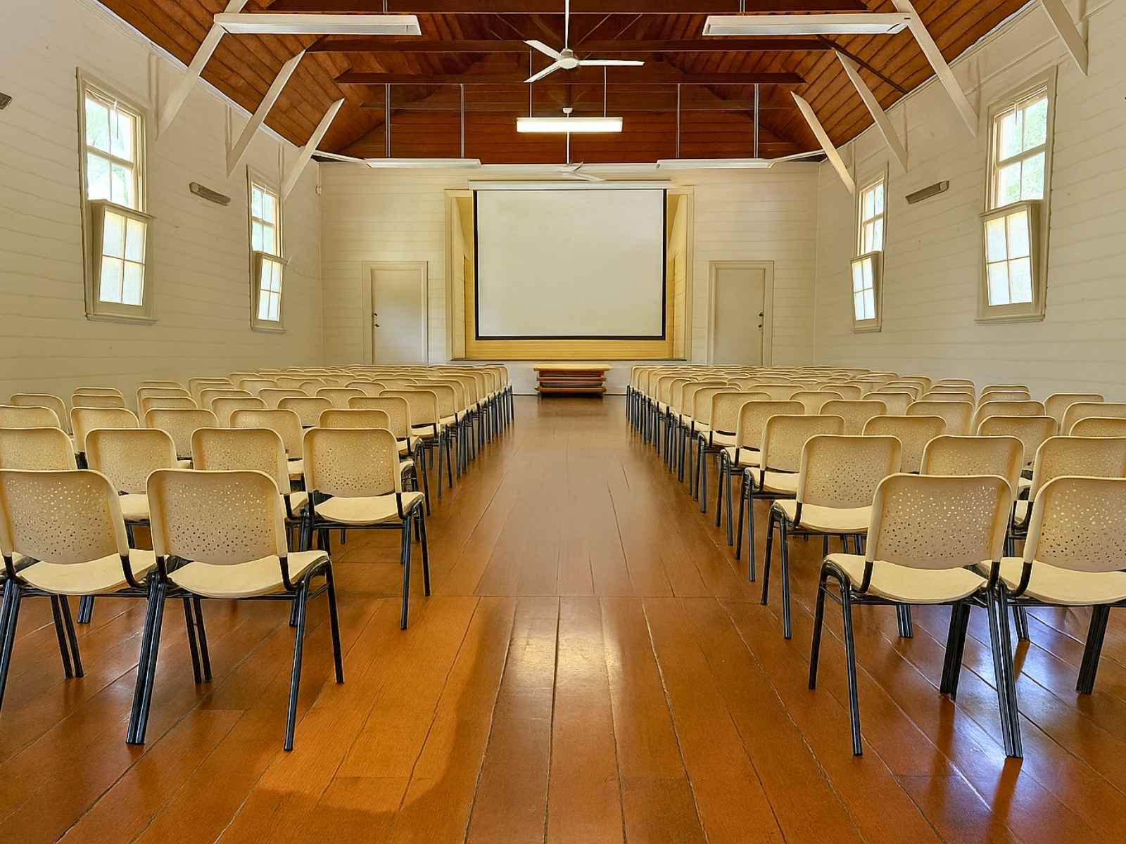 A bright and functional setup inside the historic hall, with rows of beige chairs facing a large projection screen, framed by white timber walls and a rich timber ceiling.