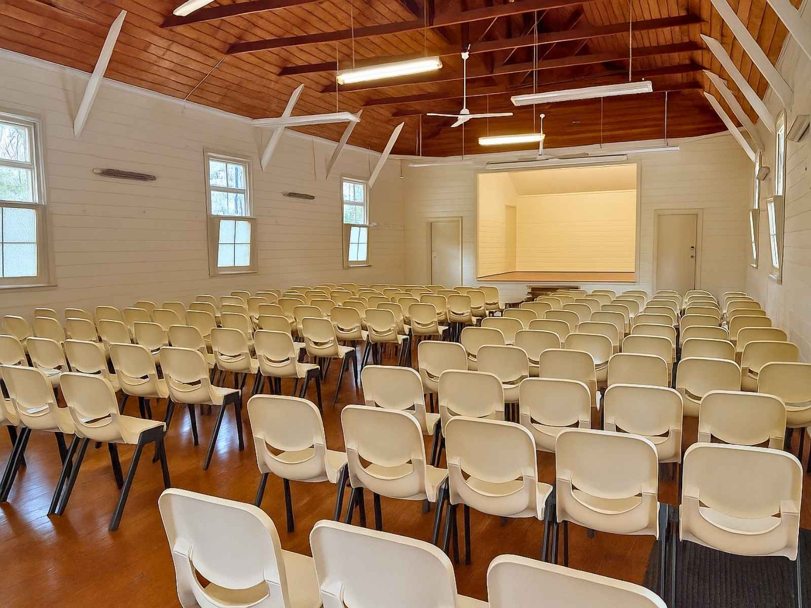 A wide-angle view showing plastic chairs neatly aligned in a classic community hall space with natural timber floors and a raised stage framed by white panelled walls.
