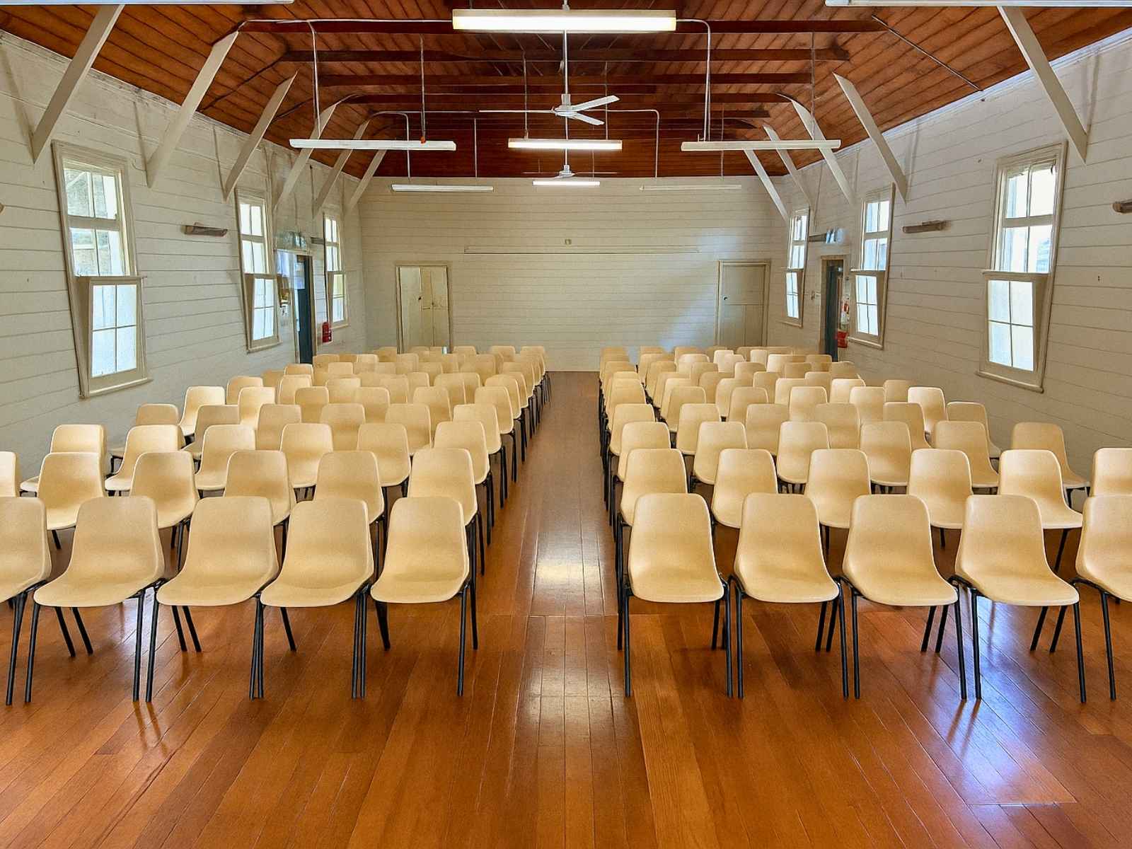 A symmetrical view down the centre aisle of Red Hill Mechanics Hall, showing cream-coloured plastic chairs, warm timber floors, and multiple windows letting in natural light.