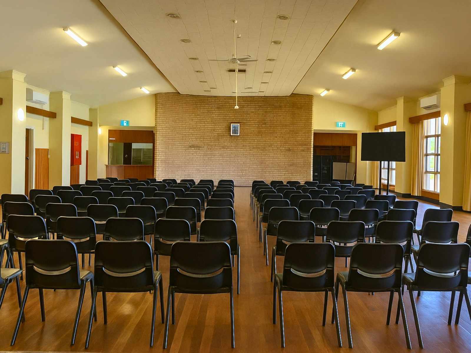 The view from the stage facing the audience, showing symmetrical windows, wall-mounted TVs, and a small serving hatch or bar to the side.