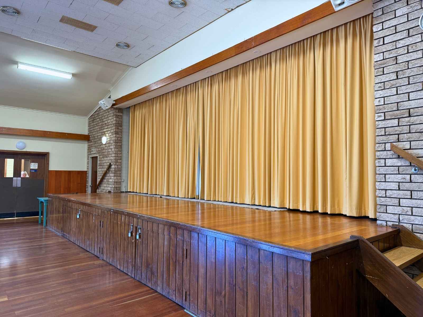 A side view of a raised timber stage inside the hall, framed by light brick walls and long mustard-yellow stage curtains.