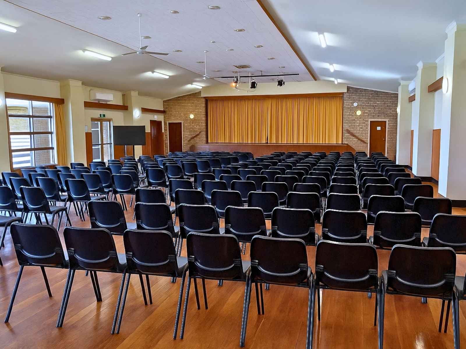A large, bright auditorium with rows of black plastic chairs on polished wooden floors facing a stage with golden curtains.