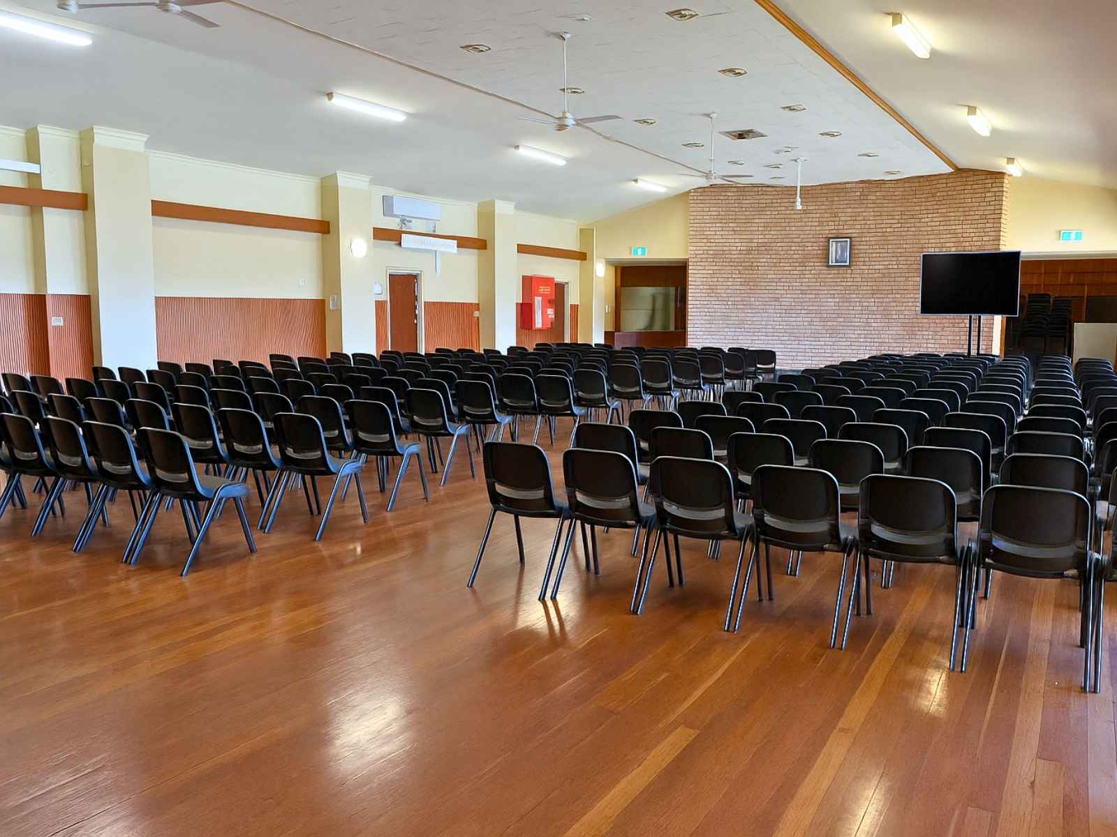 An expansive view showing the full width of the auditorium, lined with rows of black chairs, timber flooring, and a gently vaulted ceiling with fans and lights.