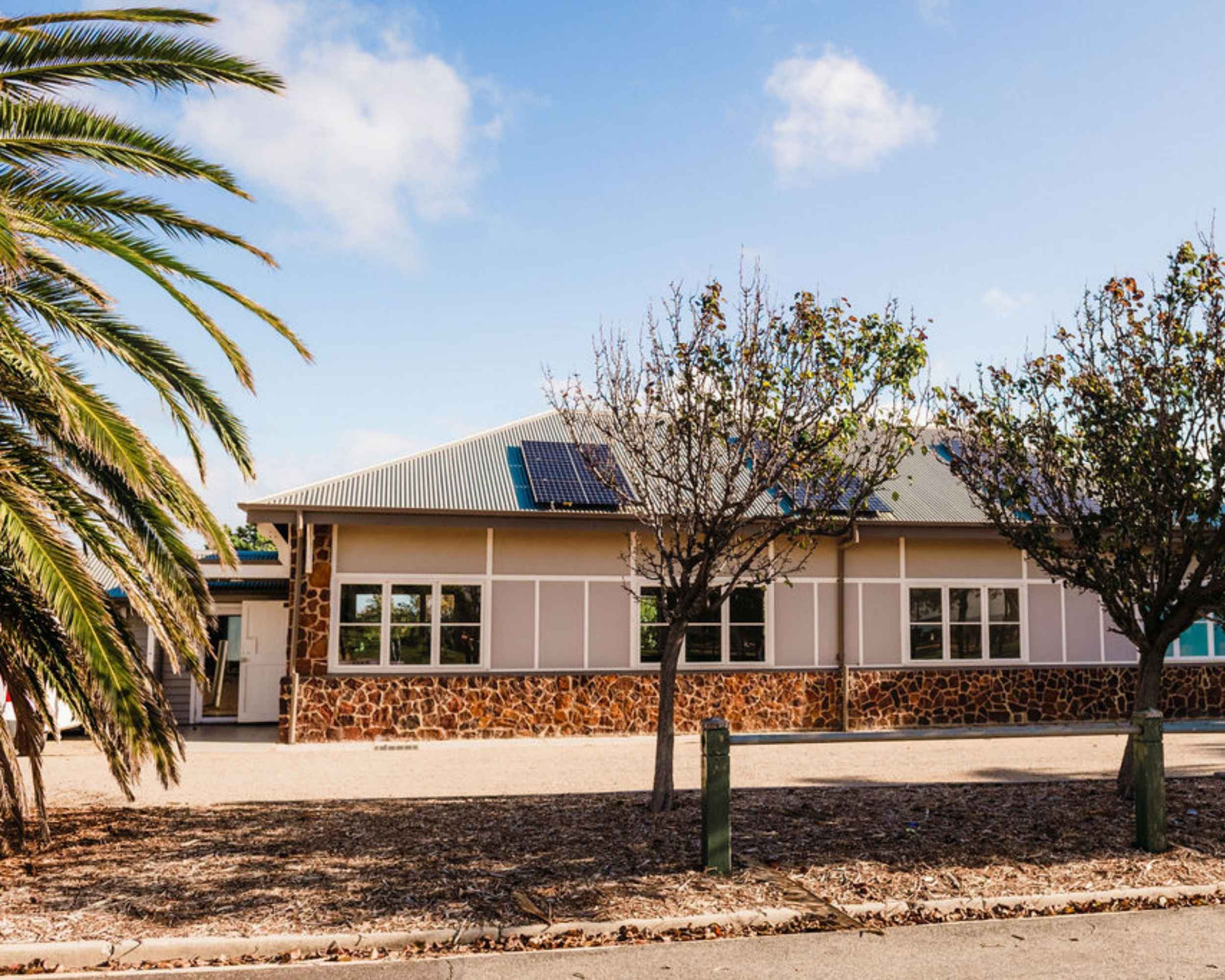 Exterior view of a community hall with cream weatherboard walls, stone accents, and solar panels on the roof, framed by palm trees and deciduous trees in the foreground.