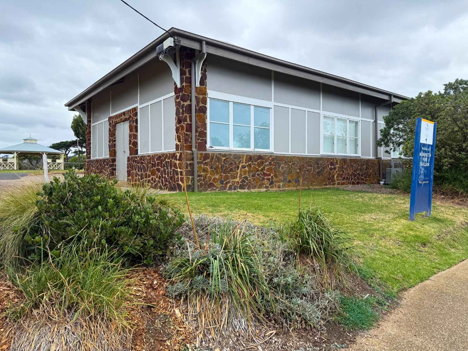 Corner view of the same hall, showing its red stone lower facade, pale grey upper panels, and a prominent sign indicating it's the Mornington Park Pavilion.