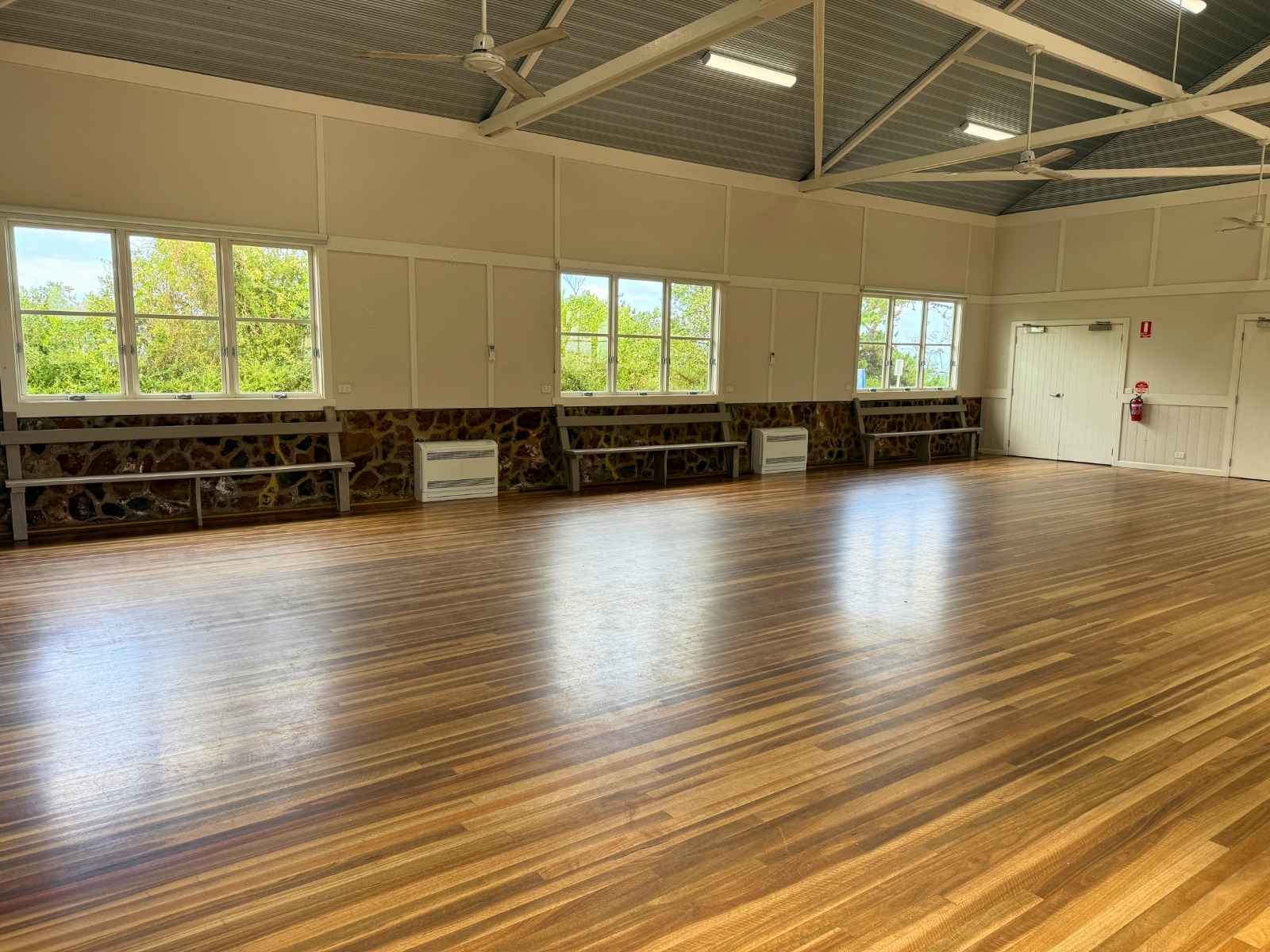 Side angle of the empty hall with exposed beams, timber floors, bench seating along the walls, and large windows looking out onto green foliage.