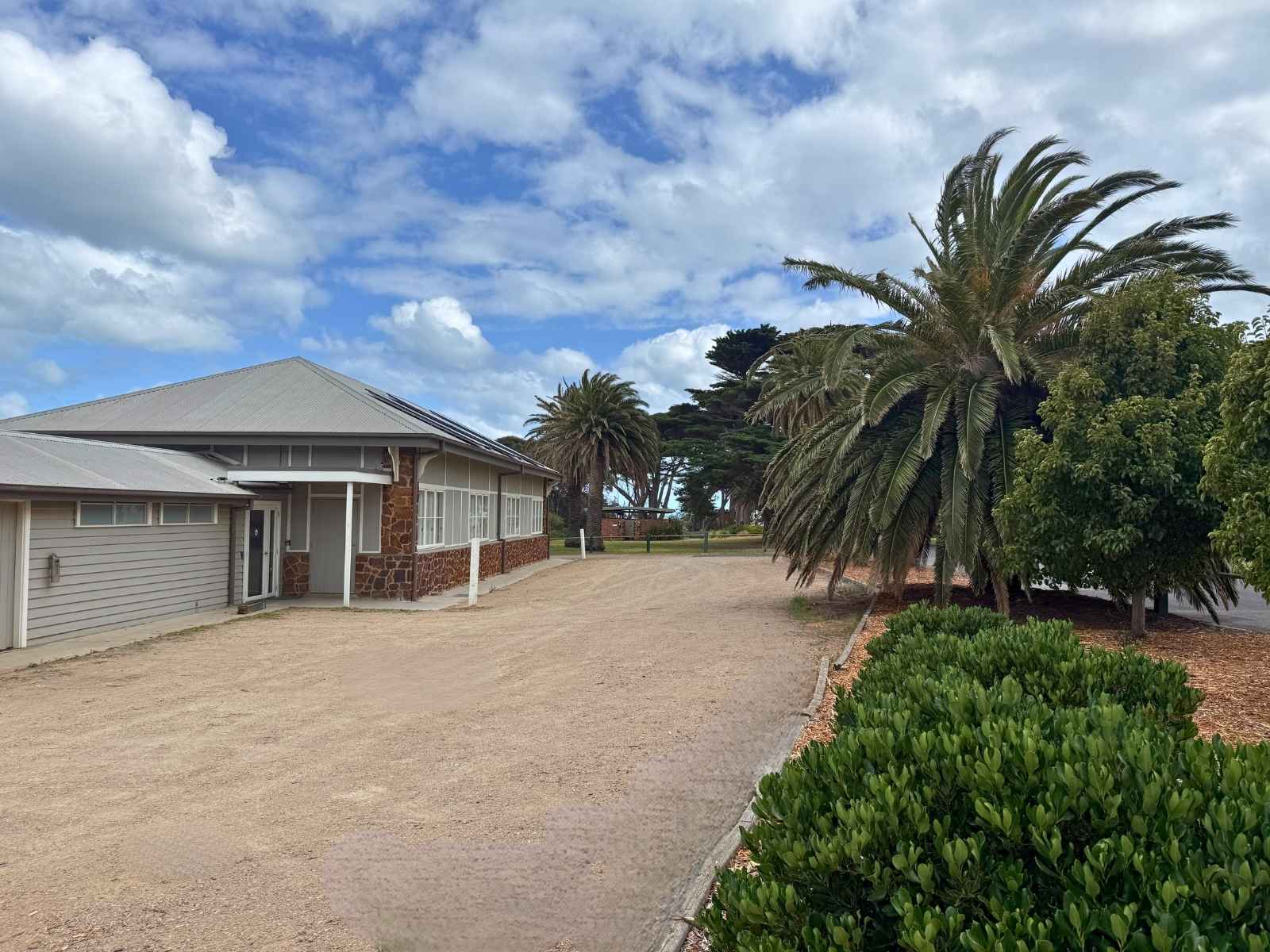 Rear view of the hall’s exterior showing the building's side entry, natural stone details, and a surrounding landscape of mature palms and coastal shrubs.