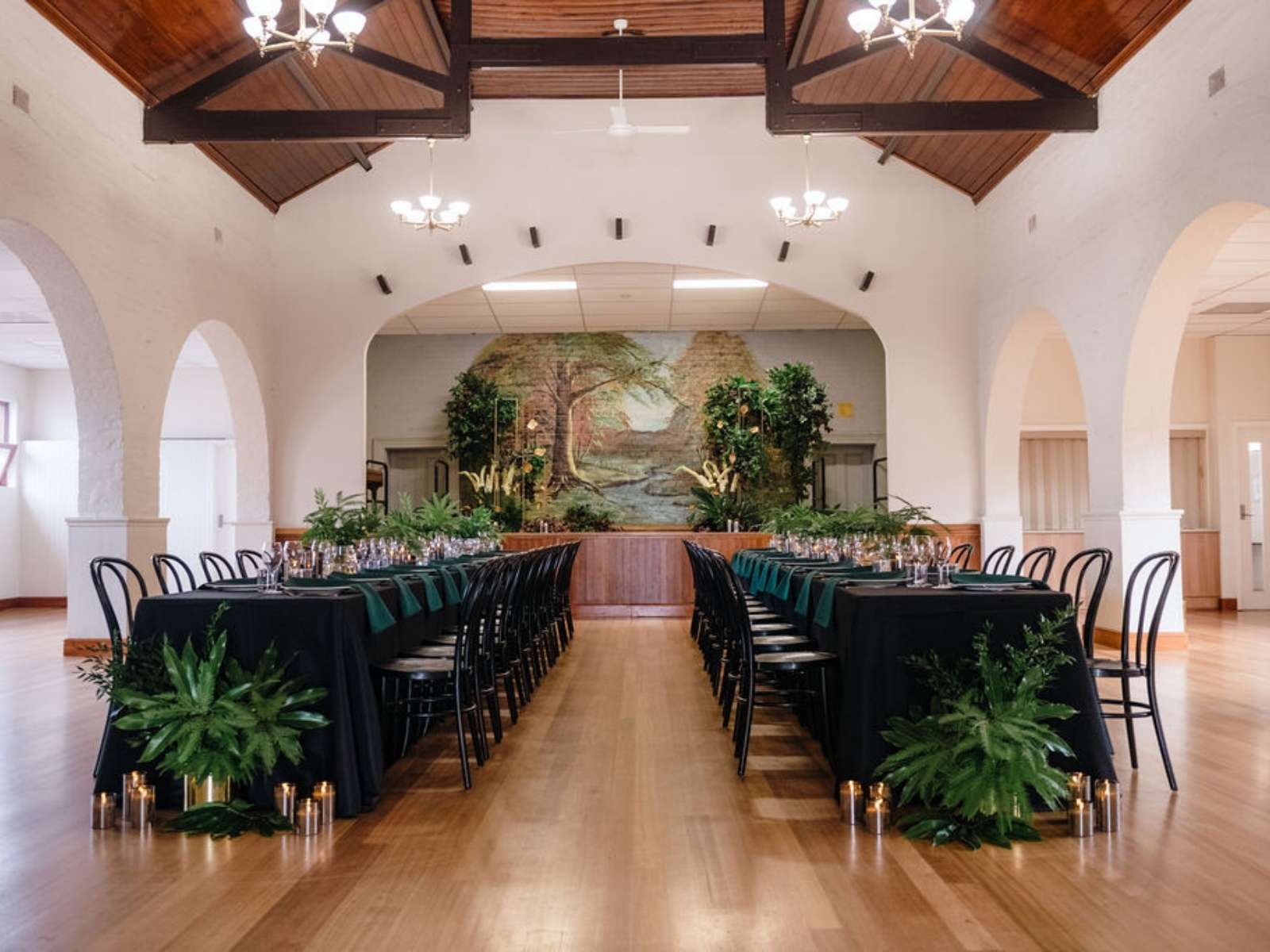 Formal dinner setup at Somerville Mechanics Hall with long black tablecloths, greenery, and stage in background.