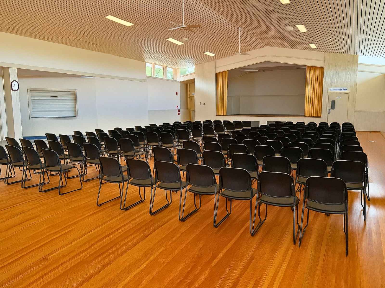 Spacious hall interior at Tyabb Community Hall with rows of black chairs facing a raised stage with closed yellow curtains.