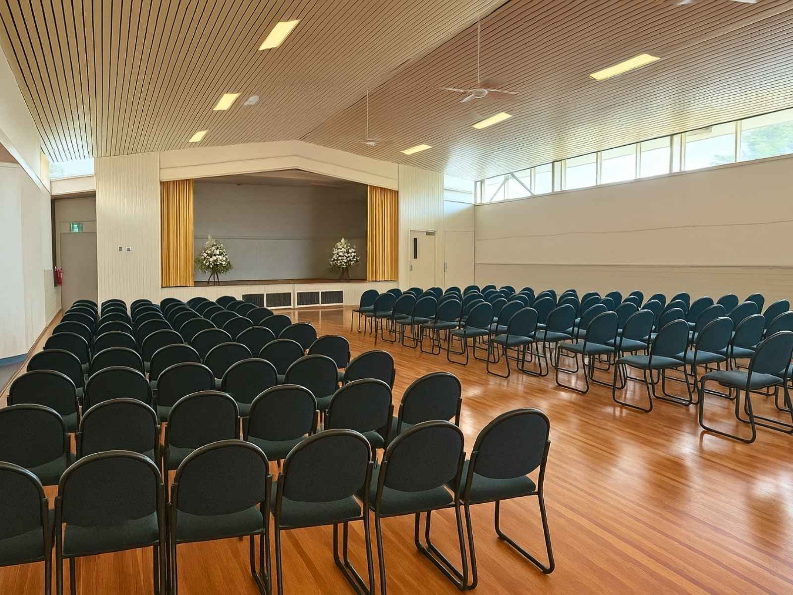 Large hall setup with dark upholstered chairs and two white floral arrangements on either side of the stage.