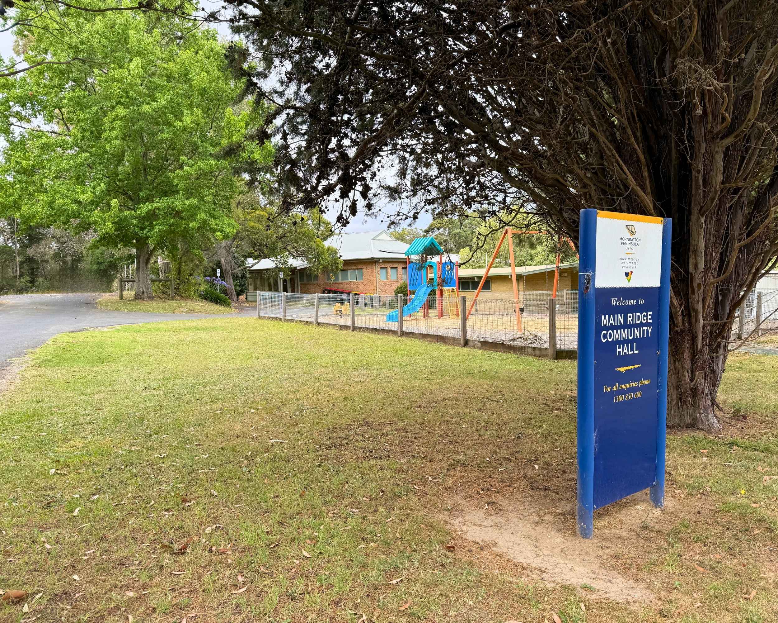 Exterior view of Main Ridge Community Hall with playground in the background and large tree in the foreground, alongside a blue welcome sign.