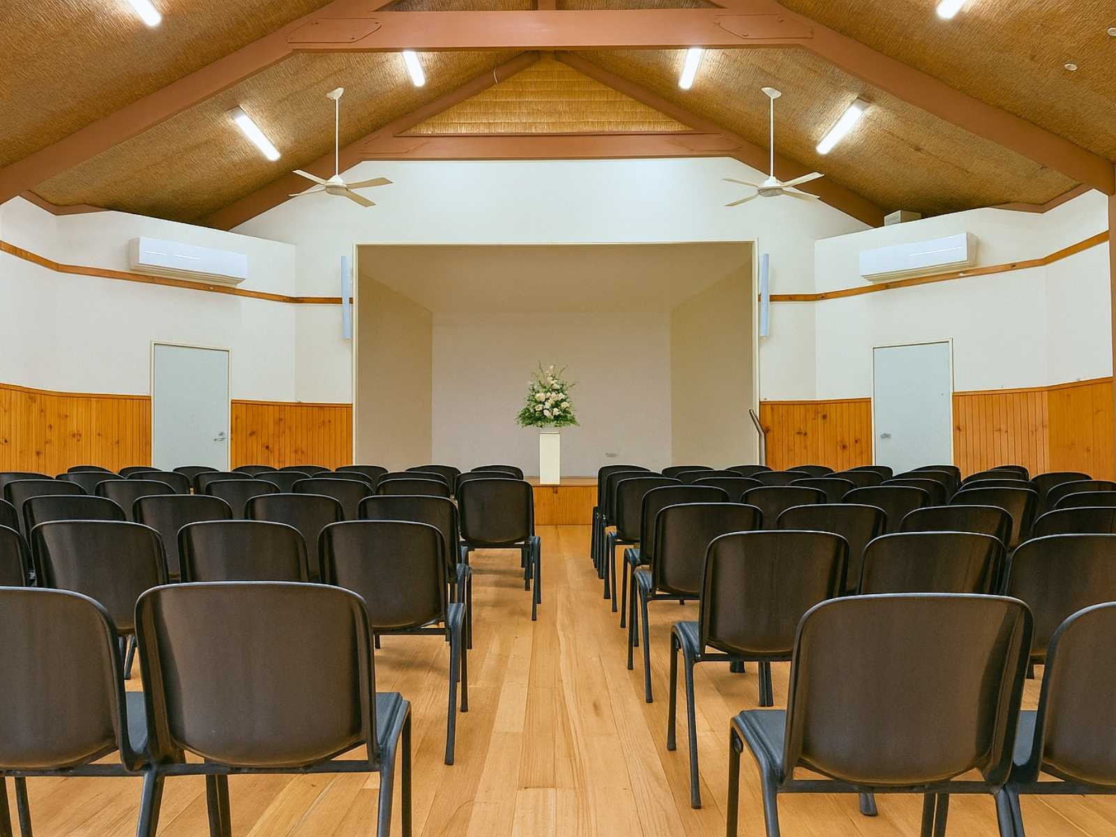 Front-facing view of the stage inside Main Ridge Community Hall, with neatly arranged black chairs and a floral arrangement on stage.
