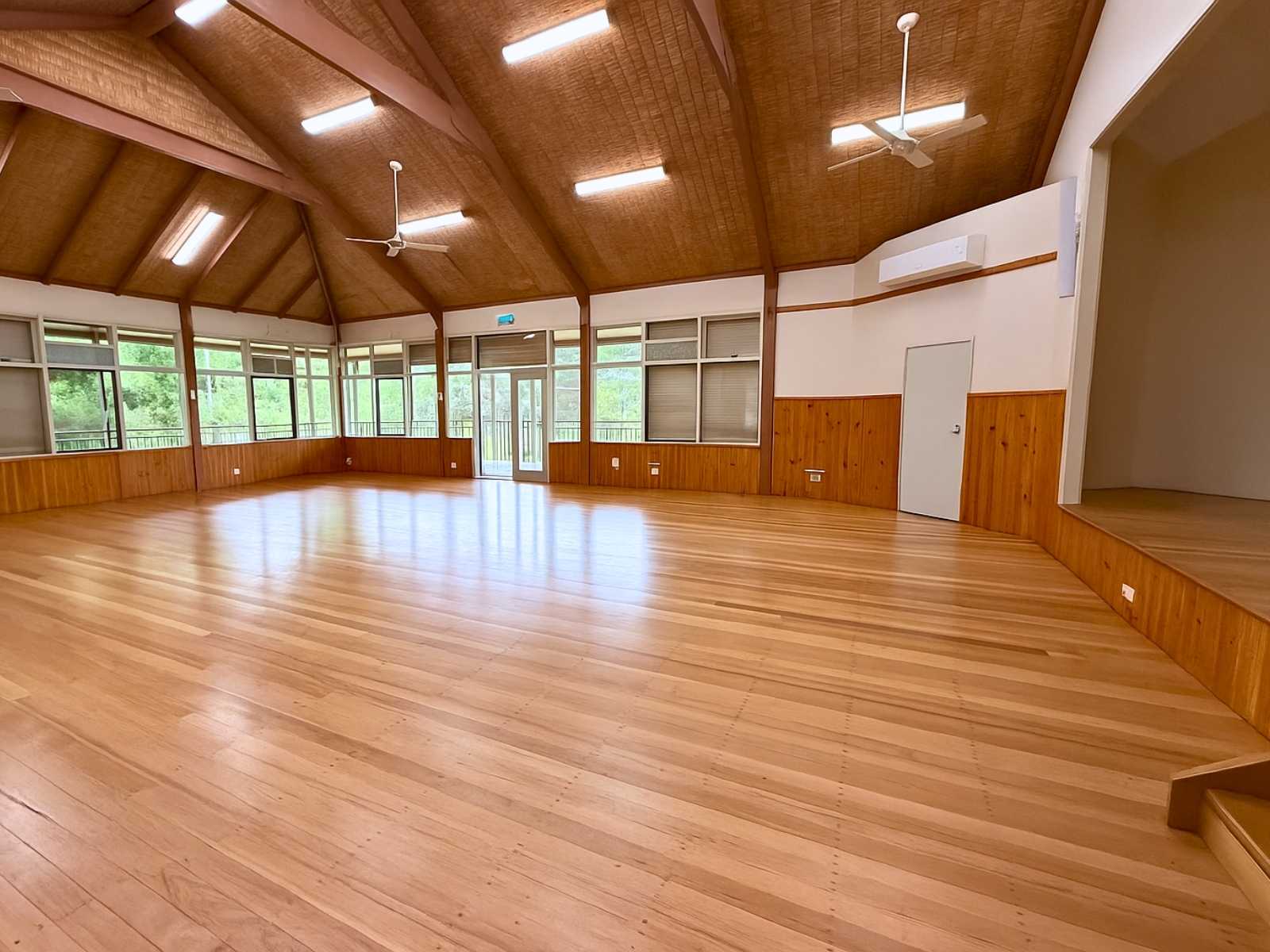Empty hall at Main Ridge Community Hall with polished timber floors, ample natural light from large windows, and a visible stage area.
