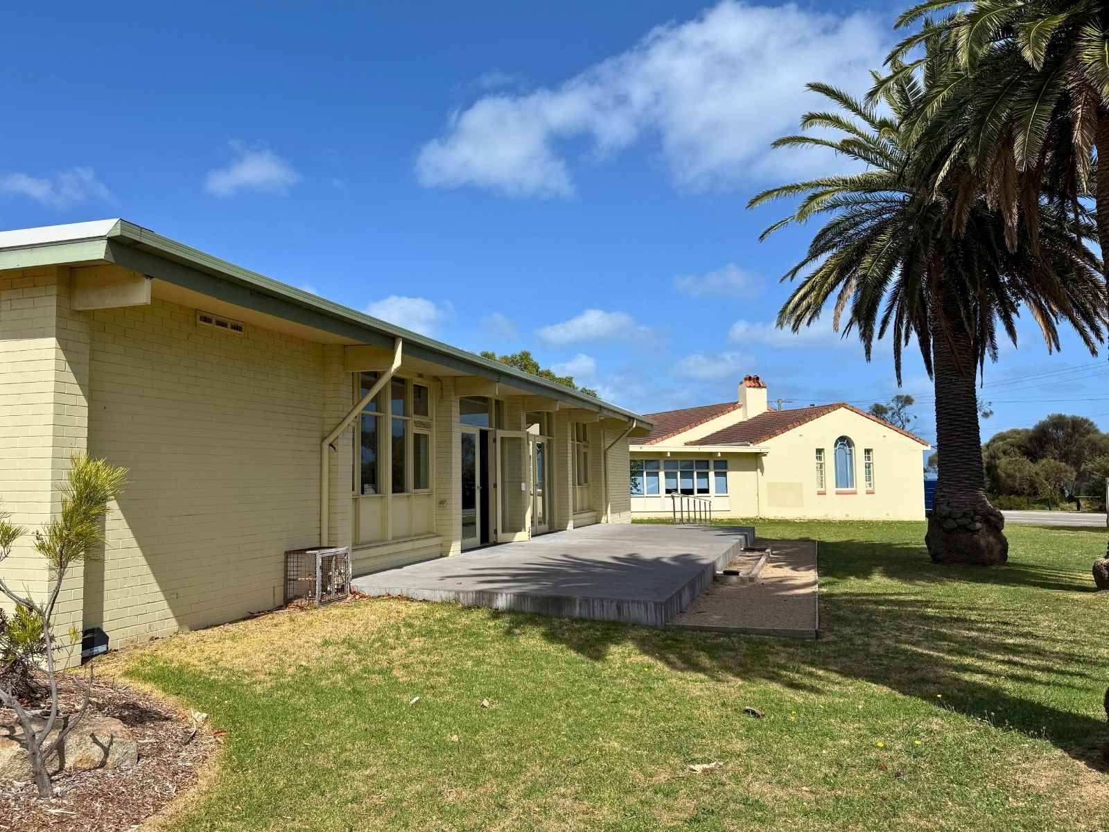 The cream brick building is surrounded by lawn and garden beds, with an adjacent hall nearby.