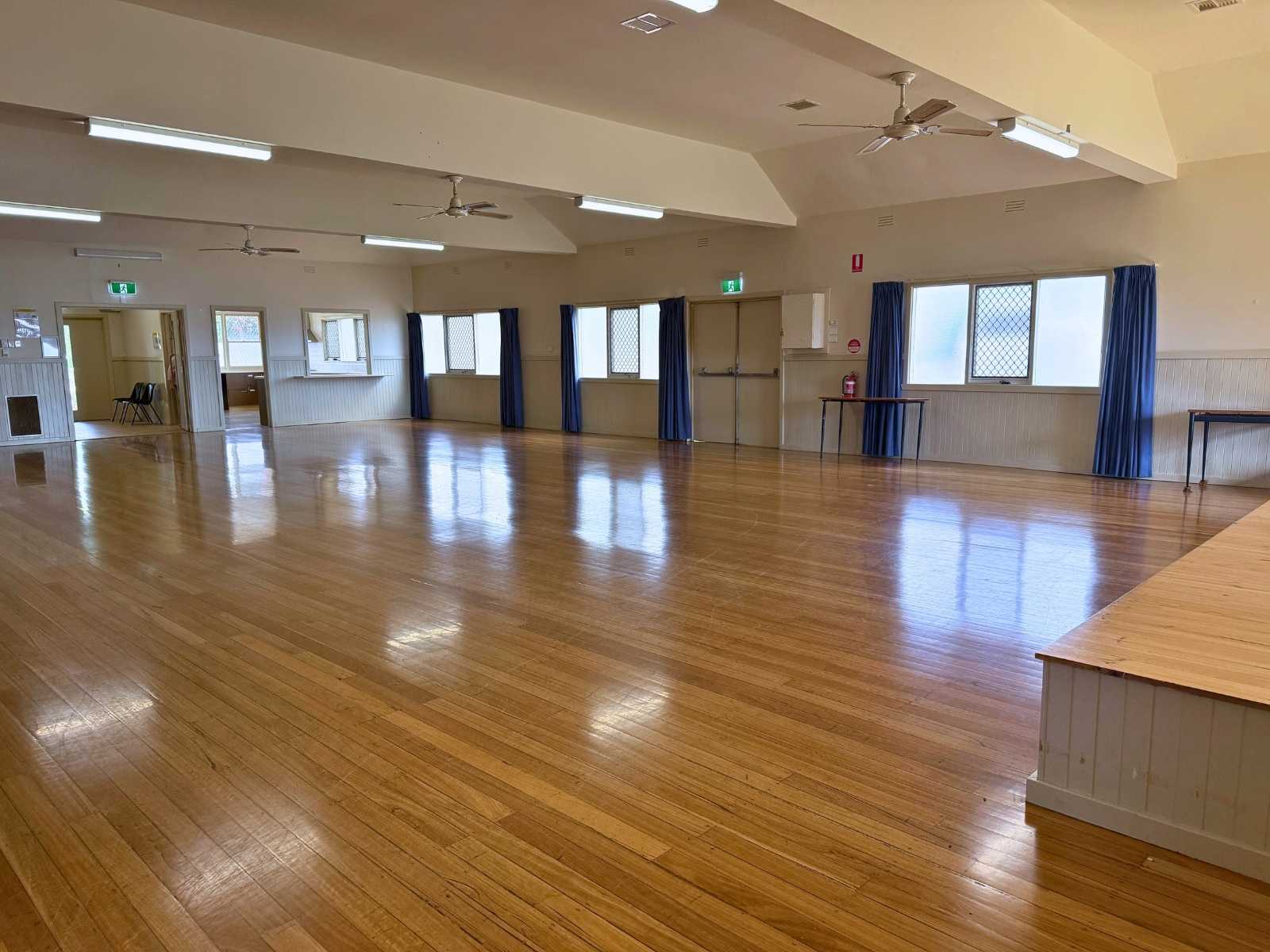 Open-plan hall with polished wooden floors, ceiling fans, and natural light from large windows.