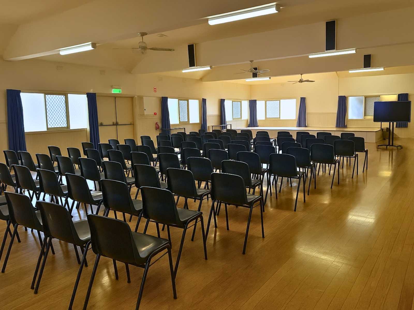 Rows of black chairs are set up inside the hall, facing a stage with natural light coming through curtained windows.