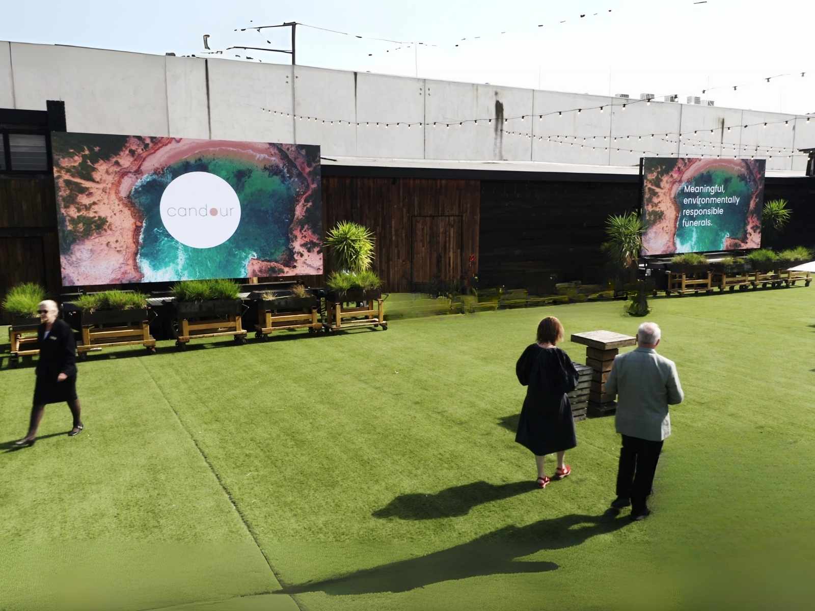 Outdoor funeral venue with large screen featuring Candour Funerals logo, surrounded by greenery and rustic wooden planters, under a partly cloudy sky.