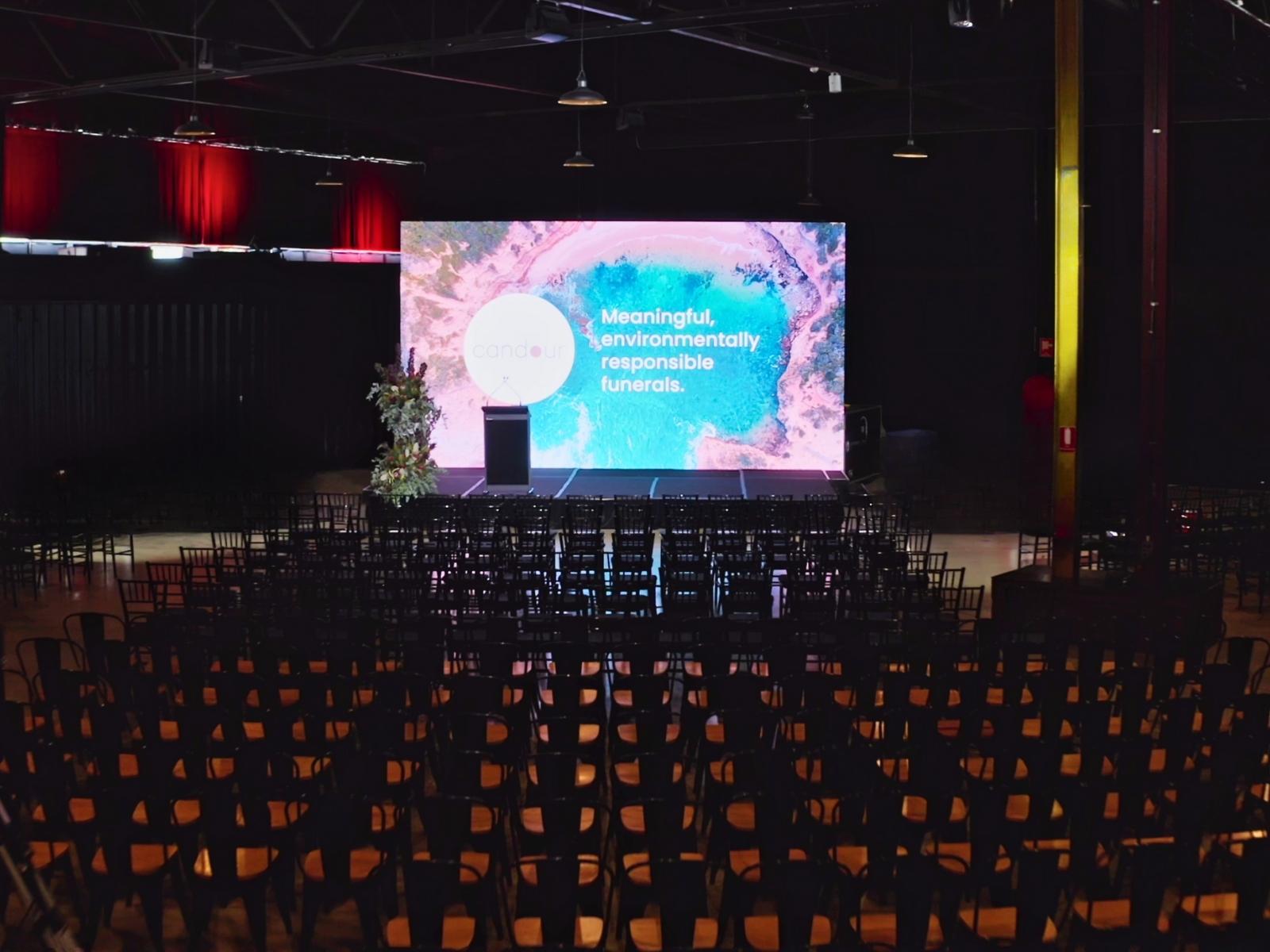 Indoor funeral venue with stage, floral arrangement, and large screen displaying "Meaningful, environmentally responsible funerals" by Candour Funerals, Melbourne.
