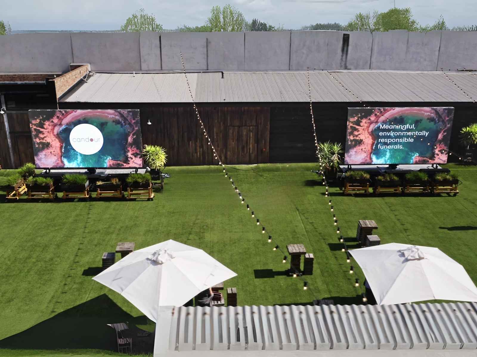 Aerial view of outdoor memorial service venue in Melbourne with twin large screens displaying Candour Funerals branding, lush green lawn, umbrellas, and string lights.