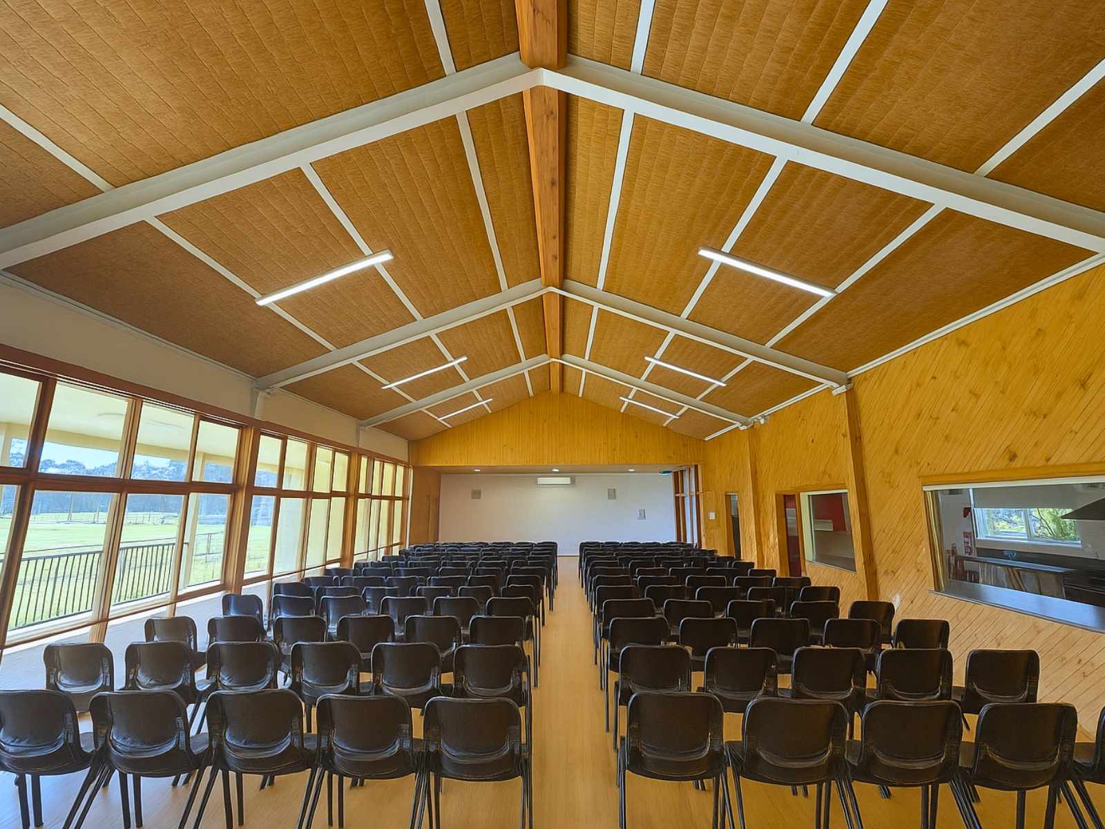 Interior view of the hall set with rows of black chairs facing a stage, with warm timber walls and ceiling.