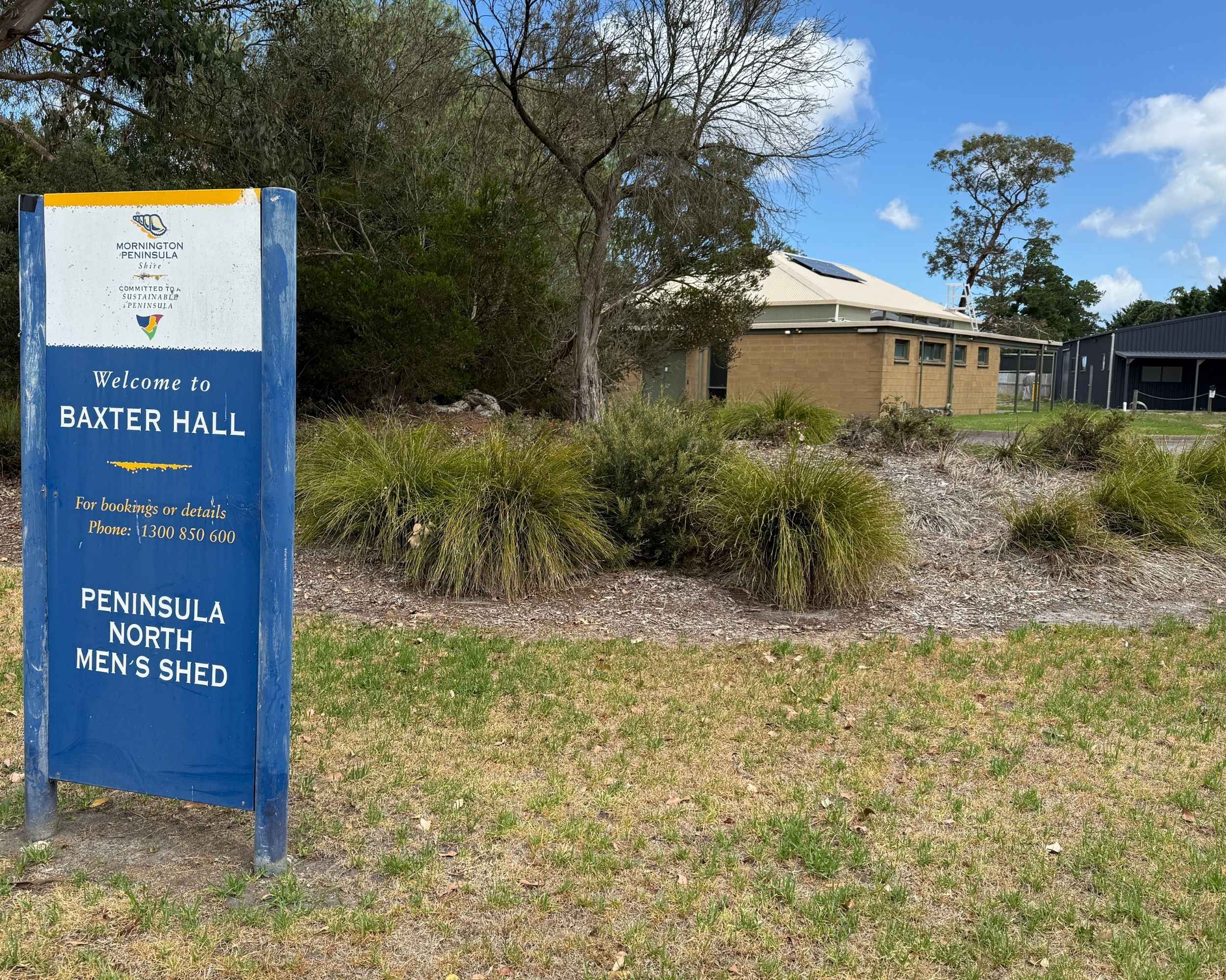 A large blue sign reading “Welcome to Baxter Hall” beside native landscaping, with the hall building visible in the background.