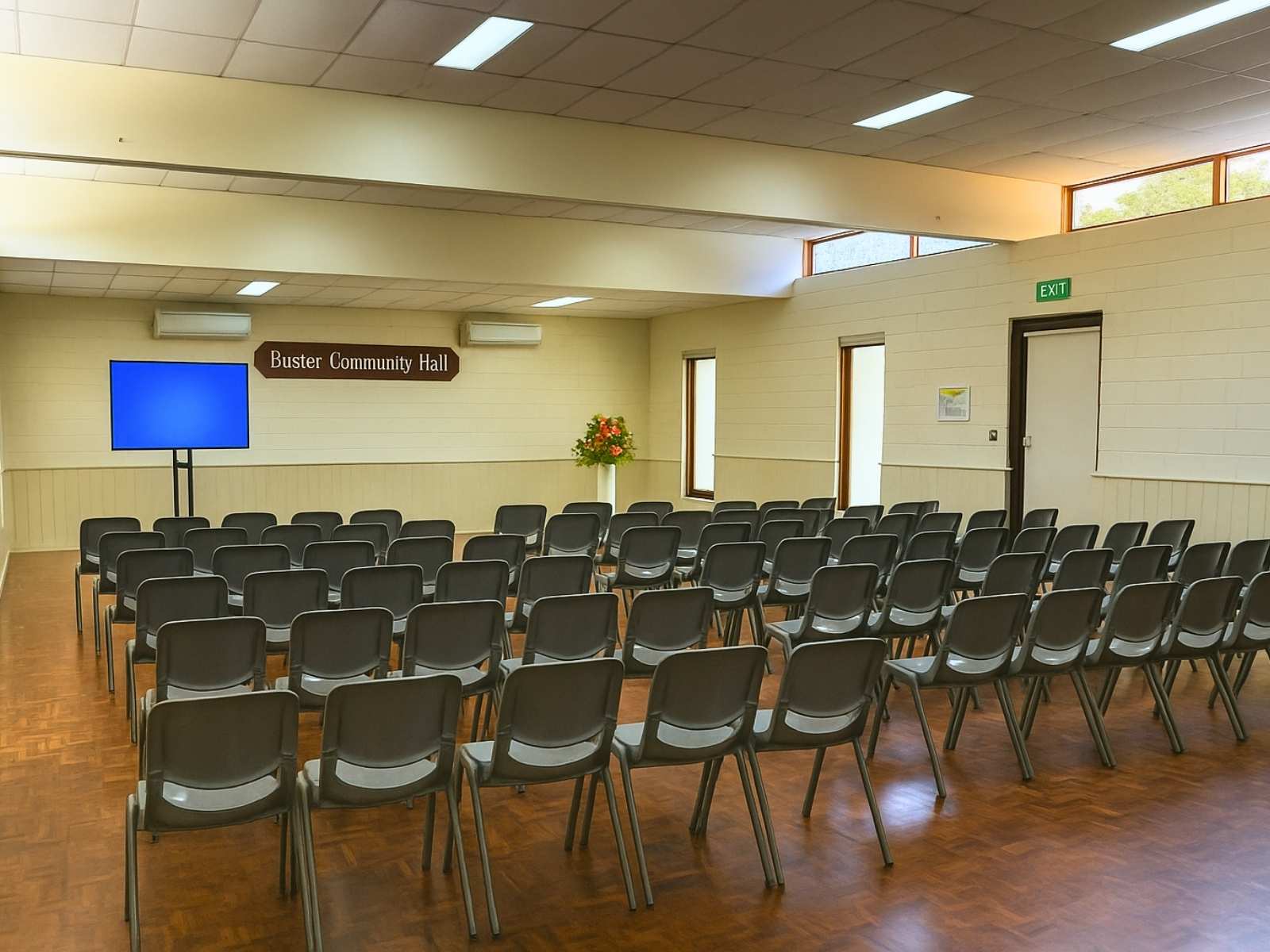 View of the hall arranged with black plastic chairs, large screen, and a floral tribute setup near the front.