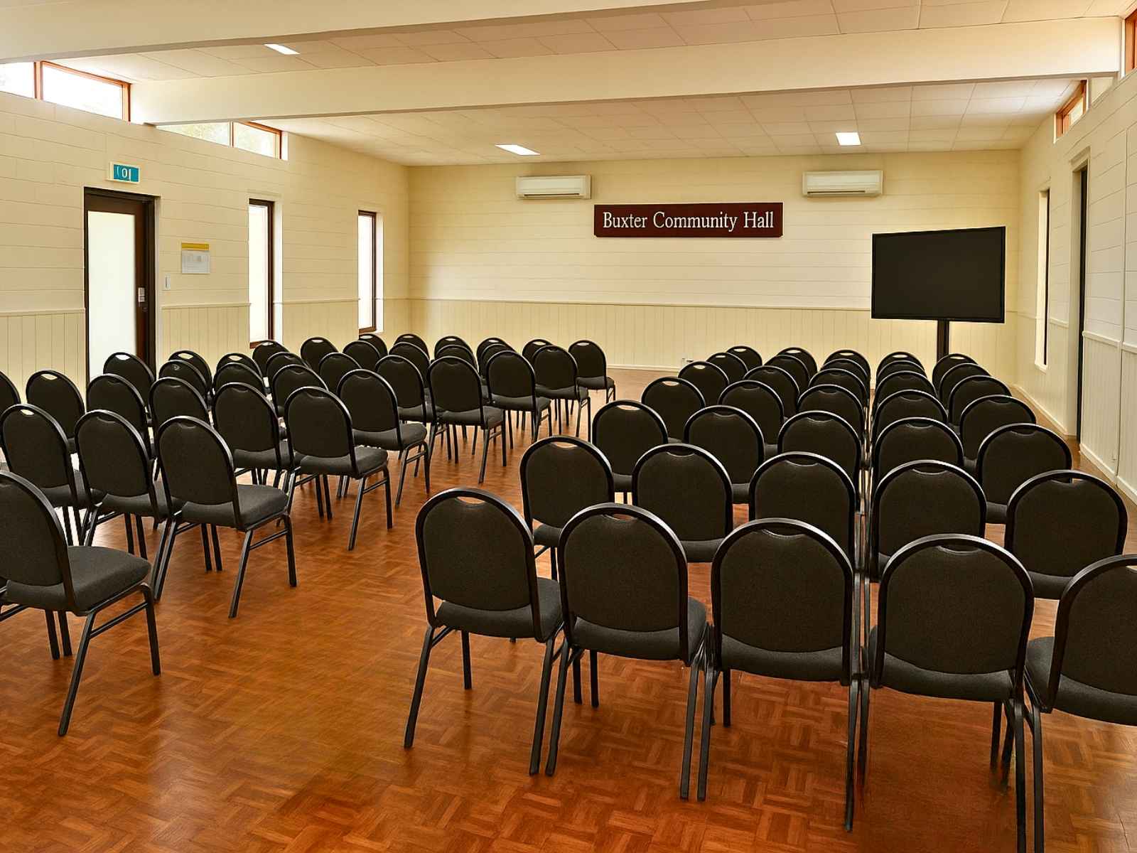 Rows of black chairs arranged theatre-style in a cream-painted hall with parquetry flooring and “Baxter Community Hall” signage at the front.
