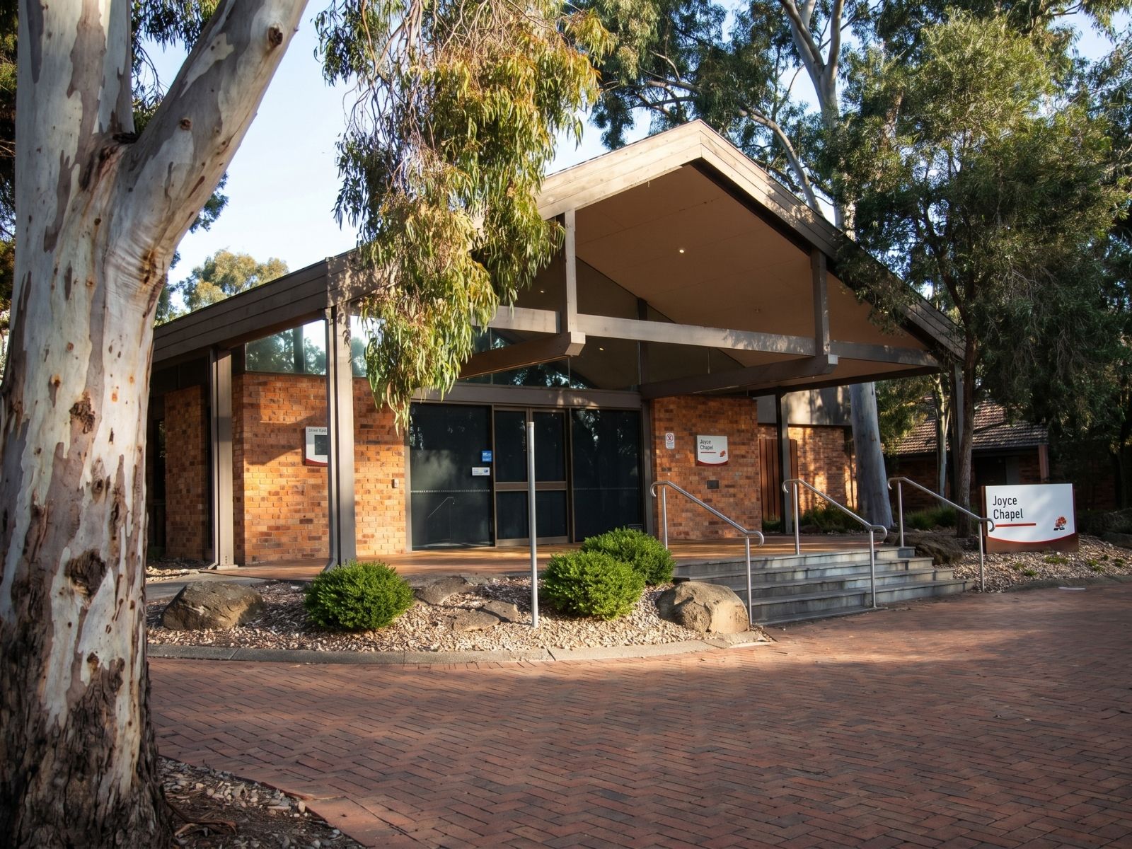 Joyce Chapel at Fawkner Memorial Park seen past a eucalyptus trunk, wide roof over the brick entry