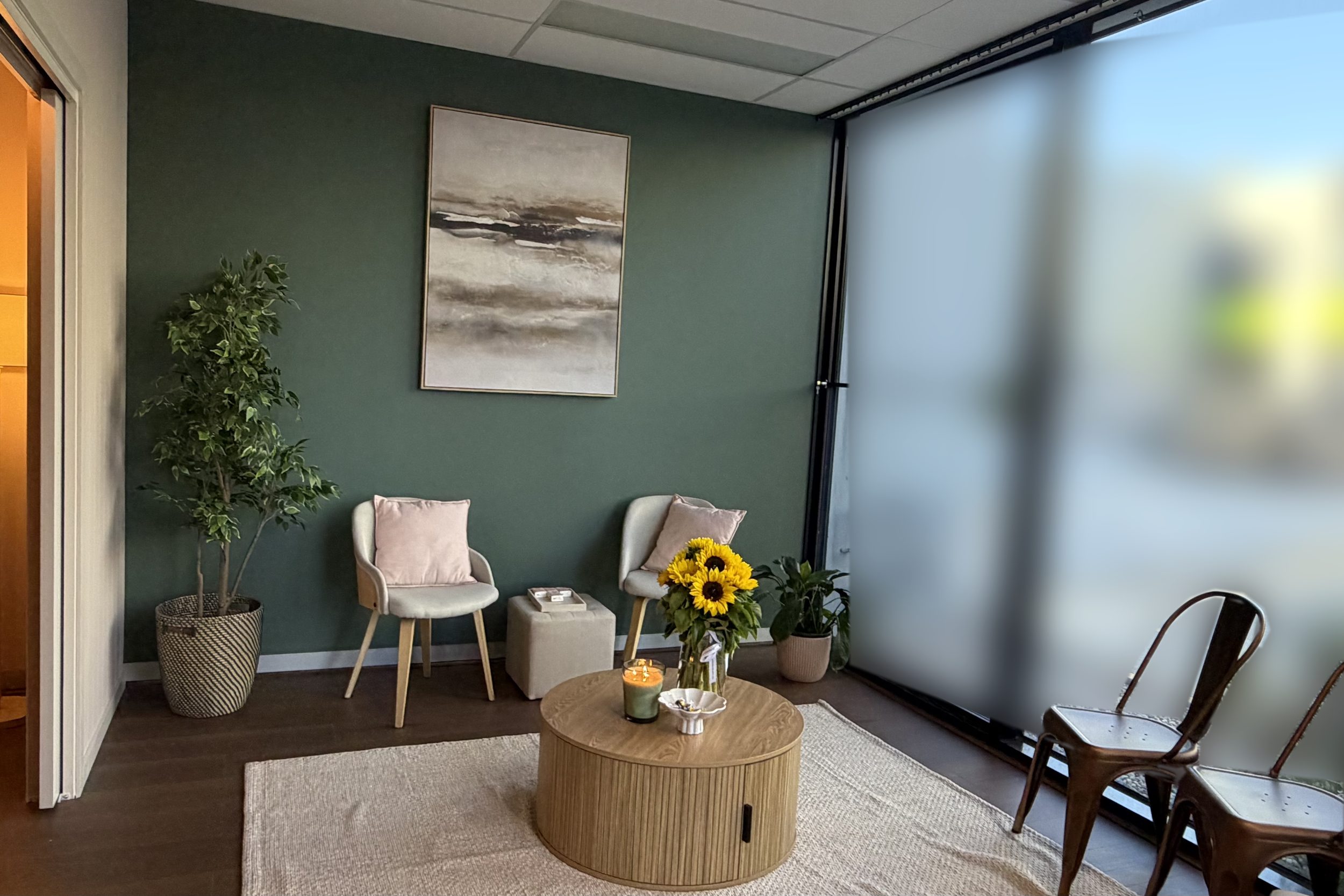 A quiet meeting room with a deep green wall, two upholstered chairs, a round timber table with sunflowers and a candle, and frosted floor-to-ceiling windows