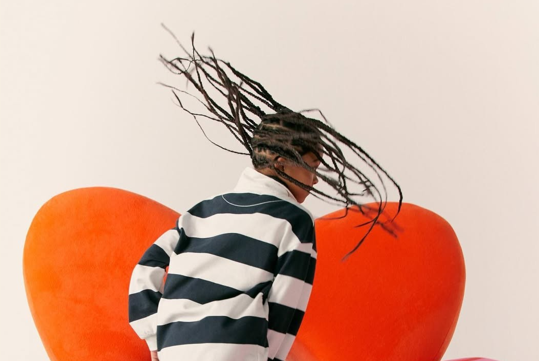 Person with braided hair flipping head back wearing a black and white striped shirt near orange cushions.