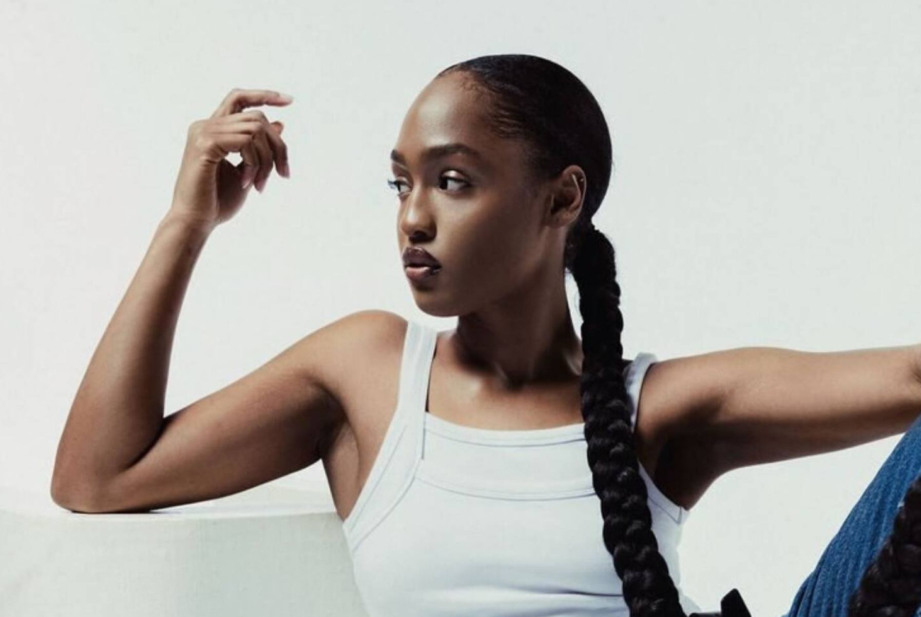 Woman with long braided hair and a white tank top looking to the side against a plain background.